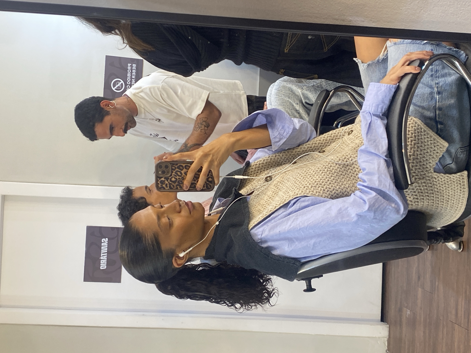 This image, taken in what appears to be a hair salon or barbershop in São Paulo, Brazil, captures a mirror selfie. The scene is rotated 90 degrees counter-clockwise.
In the foreground (bottom half of the image), a woman with slicked-back dark hair tied into a ponytail is seated in a black salon chair. She has light brown skin, her eyes are closed, and she wears white earbuds. Her attire includes a light blue button-down shirt layered with a beige knitted vest. She is holding a phone with a leopard print case, seemingly taking the mirror photo or recording.
Reflected in the mirror (top half of the image), behind the seated woman, is a man with short, dark, curly hair, a mustache, and a beard. He wears a white t-shirt, silver hoop earrings, and a necklace, and has tattoos on his forearms. He appears to be a hairdresser or barber, looking down towards the woman's head, suggesting he is attending to her hair.
The setting features white walls and a wooden floor. Visible on the reflected wall are two dark signs with reversed white text. One sign displays a crossed-out cigarette symbol, likely indicating "No Smoking," and partially visible text "BEBEB NE" (suggesting "No Drinking"). The sign below it clearly reads "SANITÁRIO" (Portuguese for "Restroom" or "Toilet"). The overall impression is one of a calm, everyday scene in a professional hair care establishment.