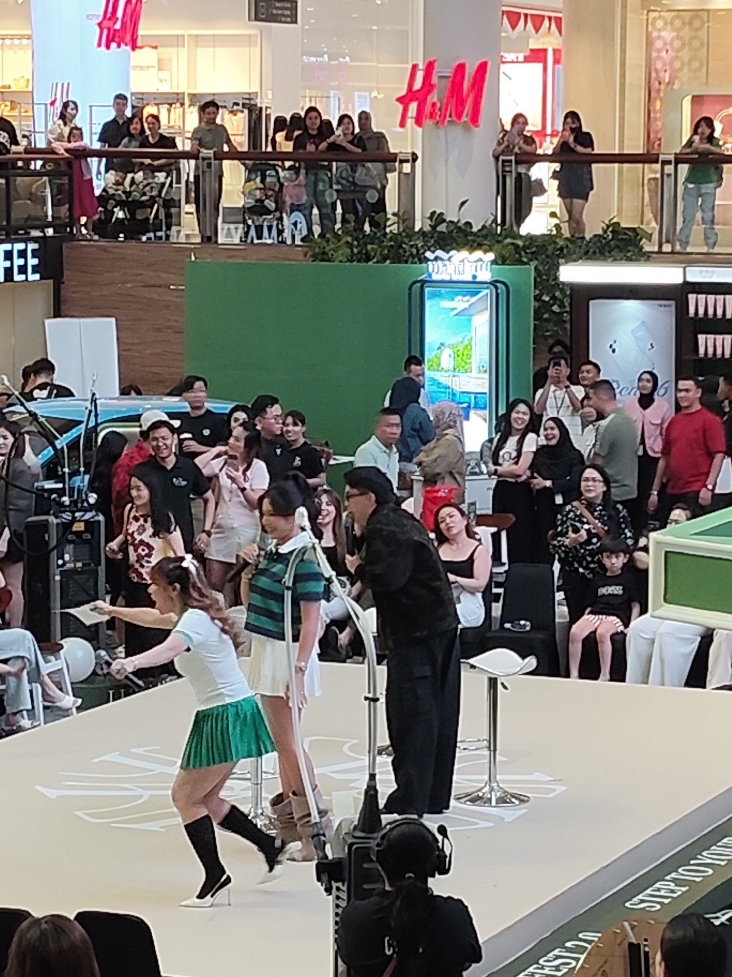 In a mall in the city of Medan, Indonesia, a group of people are gathered. In the foreground, two women are on a stage, one in a green pleated skirt and white shirt, and the other in a striped green and white shirt. The woman in the green skirt is reaching out, possibly interacting with someone off-camera. Behind them, a man in a black suit is standing. In the background, more people are observing from a balcony, some are holding up phones to take pictures or record. Stores like H&M are visible in the background. There are also display stands and what appears to be a stage setup.