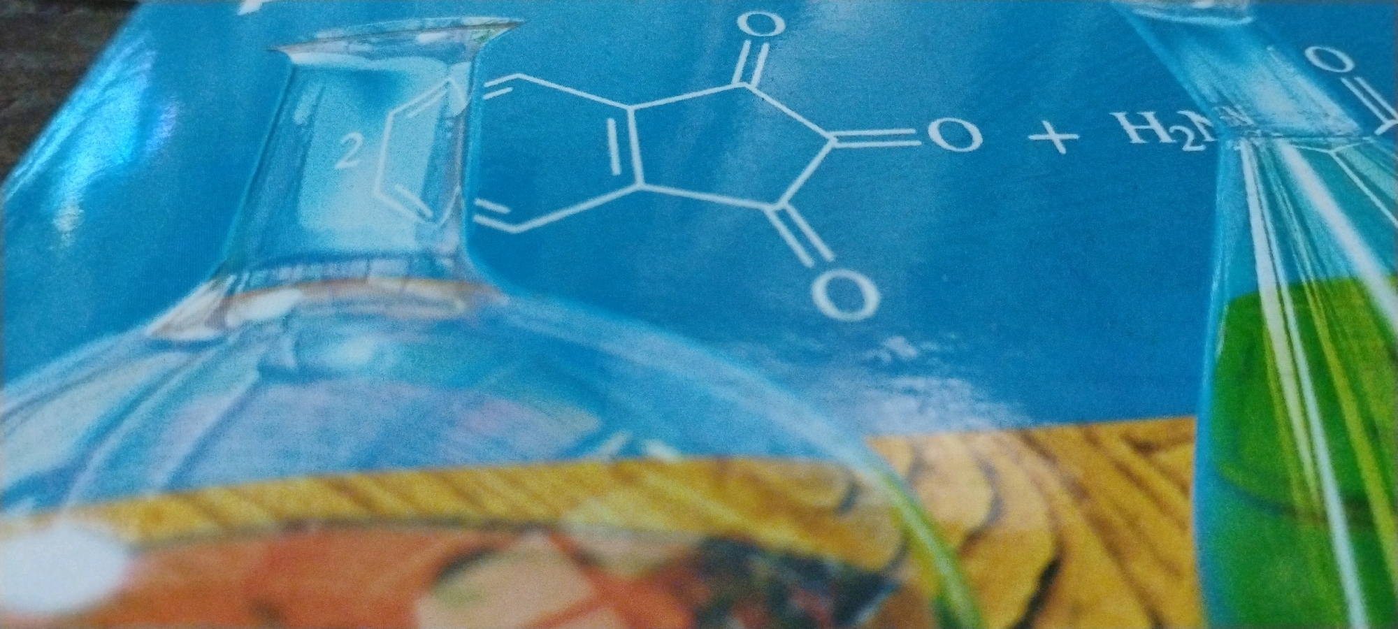The image displays a close-up view of a blue surface featuring a chemical structure diagram and parts of laboratory glassware. The chemical structure appears to be phthalic anhydride, indicated by the arrangement of carbon and oxygen atoms forming a six-membered ring fused with a five-membered ring containing two carbonyl groups. To the right of this structure, the formula "H₂O" is visible, suggesting a chemical reaction.
Dominating the foreground and left side of the image is the upper portion of a clear glass flask, possibly a volumetric flask or a distillation flask, with a flared opening. On the left side of this flask's neck, the numeral "2" is faintly visible within a hexagonal outline, which is part of the chemical structure illustration.
On the right side of the image, another piece of laboratory glassware is partially visible, appearing to be a test tube or a smaller flask containing a green liquid. The background is a solid blue color, likely representing the packaging or surface on which these elements are printed or placed. A textured, golden-yellow band runs horizontally across the lower portion of the image, beneath the glassware.
The image provides no information about the city of Jalandhar, India, or any specific location. It is a static depiction of scientific graphics and laboratory equipment.
