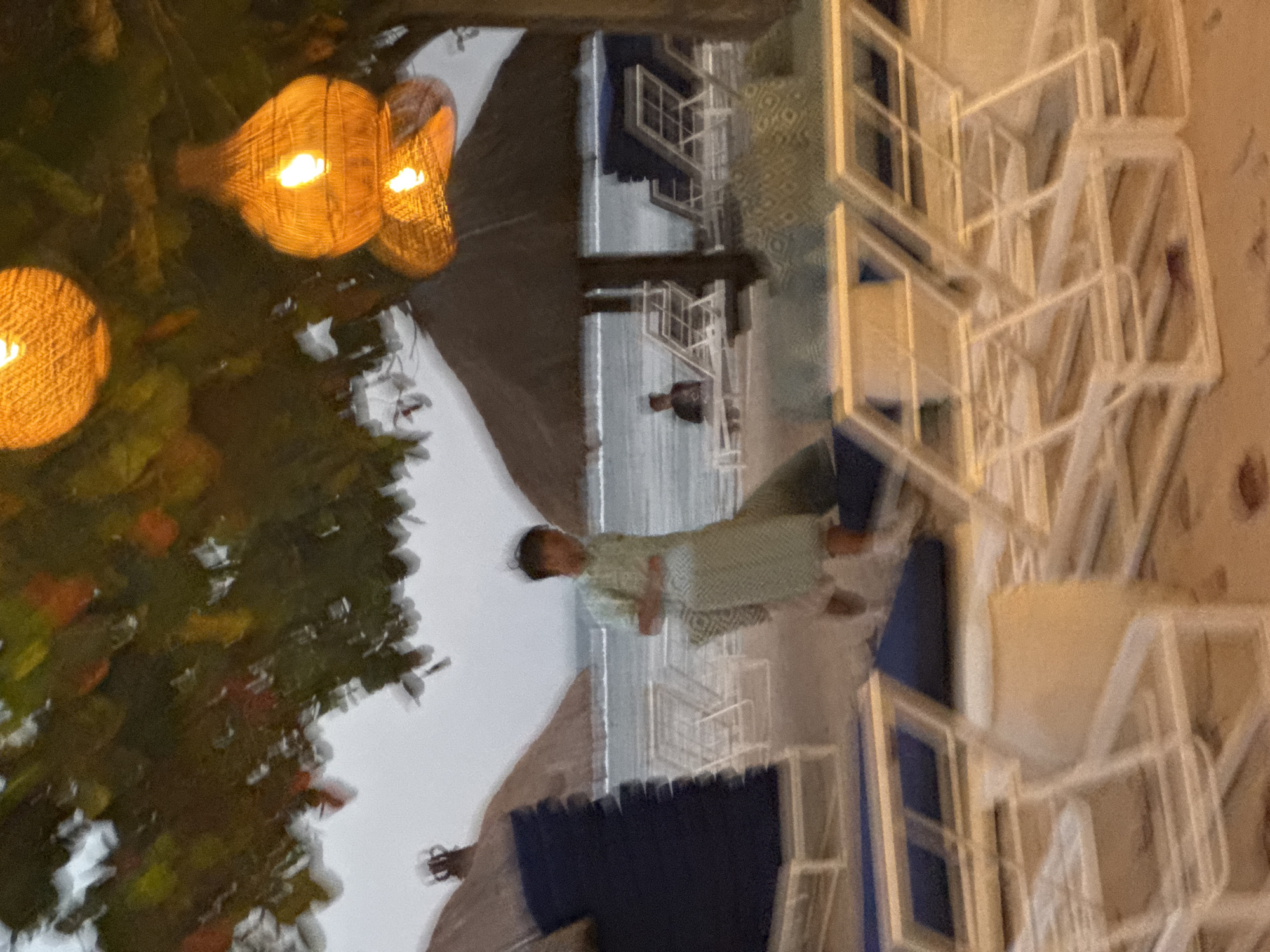 A slightly out-of-focus, medium shot captures a beach scene in Kuta Selatan, Indonesia, at dusk. Three woven, spherical lanterns, illuminated with a warm orange glow, hang from a tree branch in the foreground, casting light on the scene below. Below the lanterns, a person wearing a light-colored, patterned shirt and shorts walks to the right. In the background, several thatched-roof umbrellas are visible, along with rows of white lounge chairs. The ocean is in the distance, with a gentle wave line. A person is seated on a lounge chair under one of the umbrellas. The sand is a light brown. The sky is overcast.