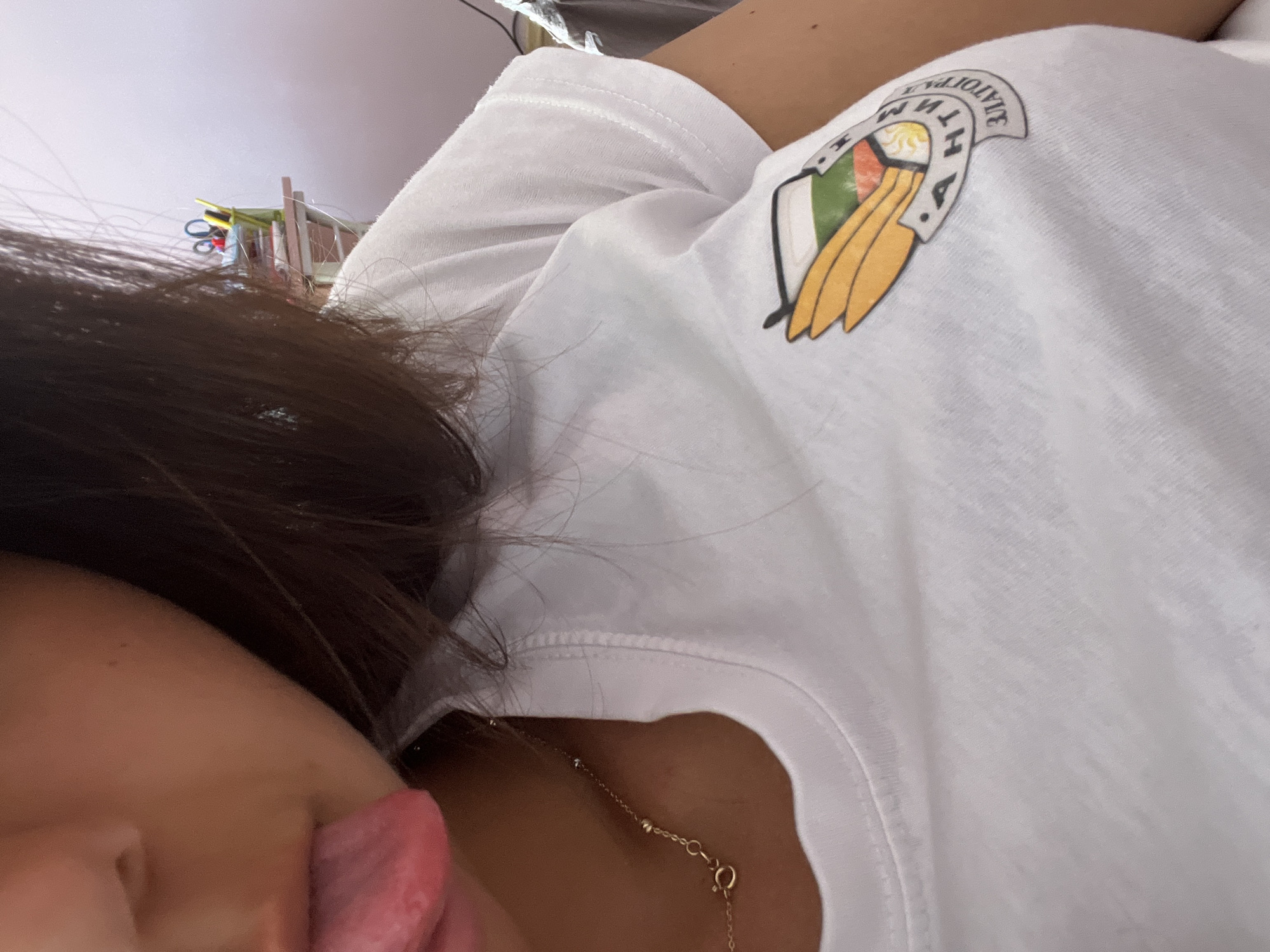 A close-up image captures a person posing informally, likely lying down, with their tongue playfully sticking out. Only a portion of their face, including the cheek, nose, and mouth area, is visible, along with their dark hair flowing around their head. They have a light-to-medium skin tone.
The person is wearing a white t-shirt that features a distinctive graphic on the shoulder. This graphic resembles a shield or emblem, incorporating a depiction of a sun, alongside sections of green, red, and white colors, and golden, feather-like or wing-like elements below. Partial text is visible on the graphic, which appears as "AHTHMI" when viewed in reverse, suggesting it's part of a longer, unidentifiable word or phrase on the emblem. A delicate gold chain necklace with small spherical beads is visible around the person's neck, resting against their skin and the neckline of the t-shirt.
The background is blurry and includes a light-colored ceiling. In the upper left area, some indistinct objects, possibly colorful cords, wires, or items on a shelf, can be vaguely discerned, indicating an indoor setting. The photograph is taken from a tilted, upward-looking perspective, reinforcing the casual and relaxed atmosphere of the pose.