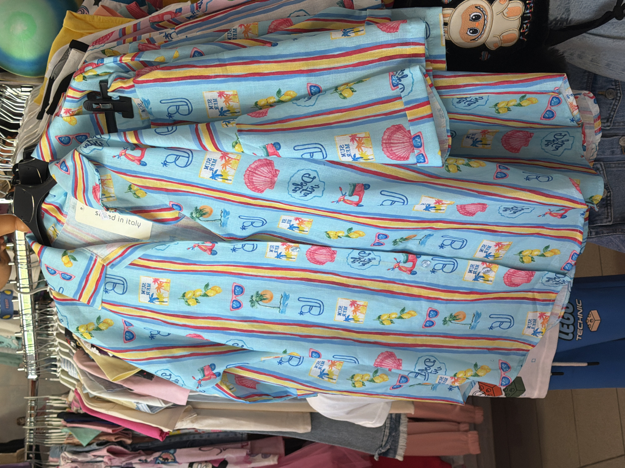 A light blue short-sleeved button-down shirt with a pattern of colorful vertical stripes and various summer-themed icons hangs on a clothing rack. The icons include lemons, sunglasses, palm trees, seashells, and scooters, interspersed with text that appears to read "SUN MER" and "Dolce Vita." A tag on the shirt reads "Made in Italy." The shirt is part of a display of clothing in what appears to be a retail setting. Other garments, including pink, white, and yellow tops, as well as blue jeans, are visible on the rack behind and below the featured shirt. The background suggests a clothing store environment with multiple racks and garments. There are no people clearly visible in the image. The location is identified as Ravno pole, Bulgaria, within a retail store.