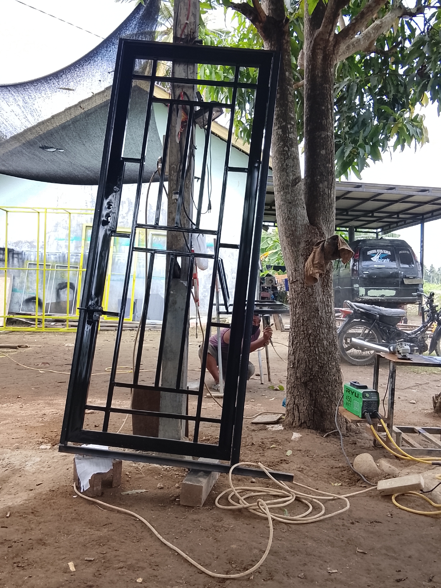 The image depicts an outdoor workshop scene in Mee, Indonesia. A large, black metal gate frame is propped up by concrete blocks, leaning against a wooden pole. To the right of the gate, a tree with rough bark stands tall. In the background, a black van and a motorcycle are parked under a corrugated metal roof structure. A person, wearing a purple shirt and a dark mask, is crouched down, appearing to be welding or working on metal. A green welding machine is visible on a wooden plank attached to the tree. The ground is dirt and sand, with yellow and white electrical cords running across it. Further back, a yellow metal frame structure with glass panels is visible. The overall impression is of an active, open-air fabrication or repair yard.