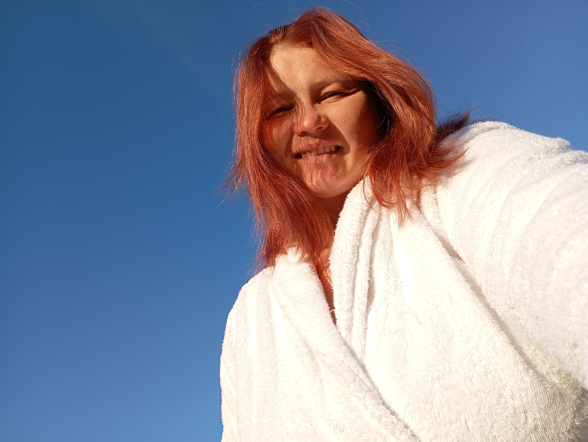 A person with reddish-brown hair is seen from a low angle, wearing a fluffy white robe, against a clear blue sky. The person is looking up and slightly to the right, with their mouth slightly open in a grimace, revealing their teeth. Sunlight casts a shadow across the left side of their face. The image appears to be a selfie taken outdoors. There is no visual information within the image to suggest a specific location in Fokino, Russia.