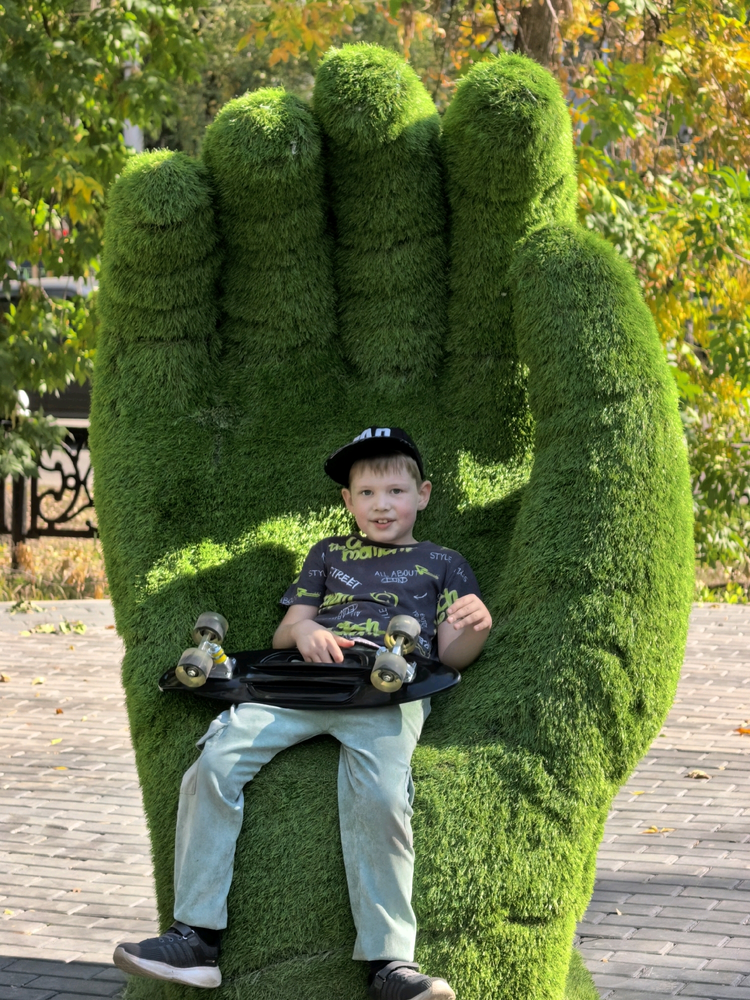 In Sterlitamak, Russia, a young boy is seated on a large, green, artificial grass sculpture shaped like a fist. He is wearing a black baseball cap, a dark t-shirt with a graphic print, and light blue pants. The boy is holding a black skateboard across his lap. The background consists of trees with some autumn foliage, suggesting the season. The ground is paved with rectangular bricks.