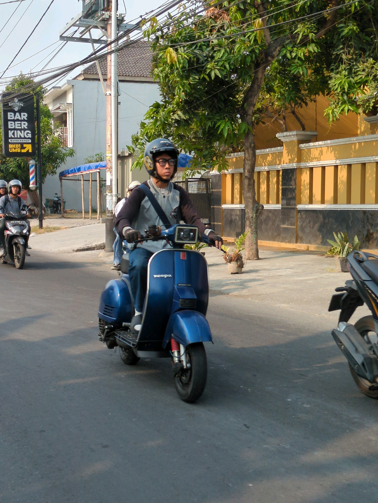 In Surakarta, Indonesia, on a paved street bathed in daylight, a young male wearing a helmet and glasses is riding a blue Vespa scooter. He is dressed in a grey and black long-sleeved shirt, blue jeans, and gloves. The Vespa has a white seat and red shock absorbers. To his left, another male is riding a black motorcycle, also wearing a helmet. Further in the background, two more individuals on motorcycles are visible. The environment features a yellow building with a decorative fence, trees, utility poles with numerous wires, and a barber shop sign that reads "BARBERKING BARBERSHOP BK01 UMS." The street is marked with some faint shadows.