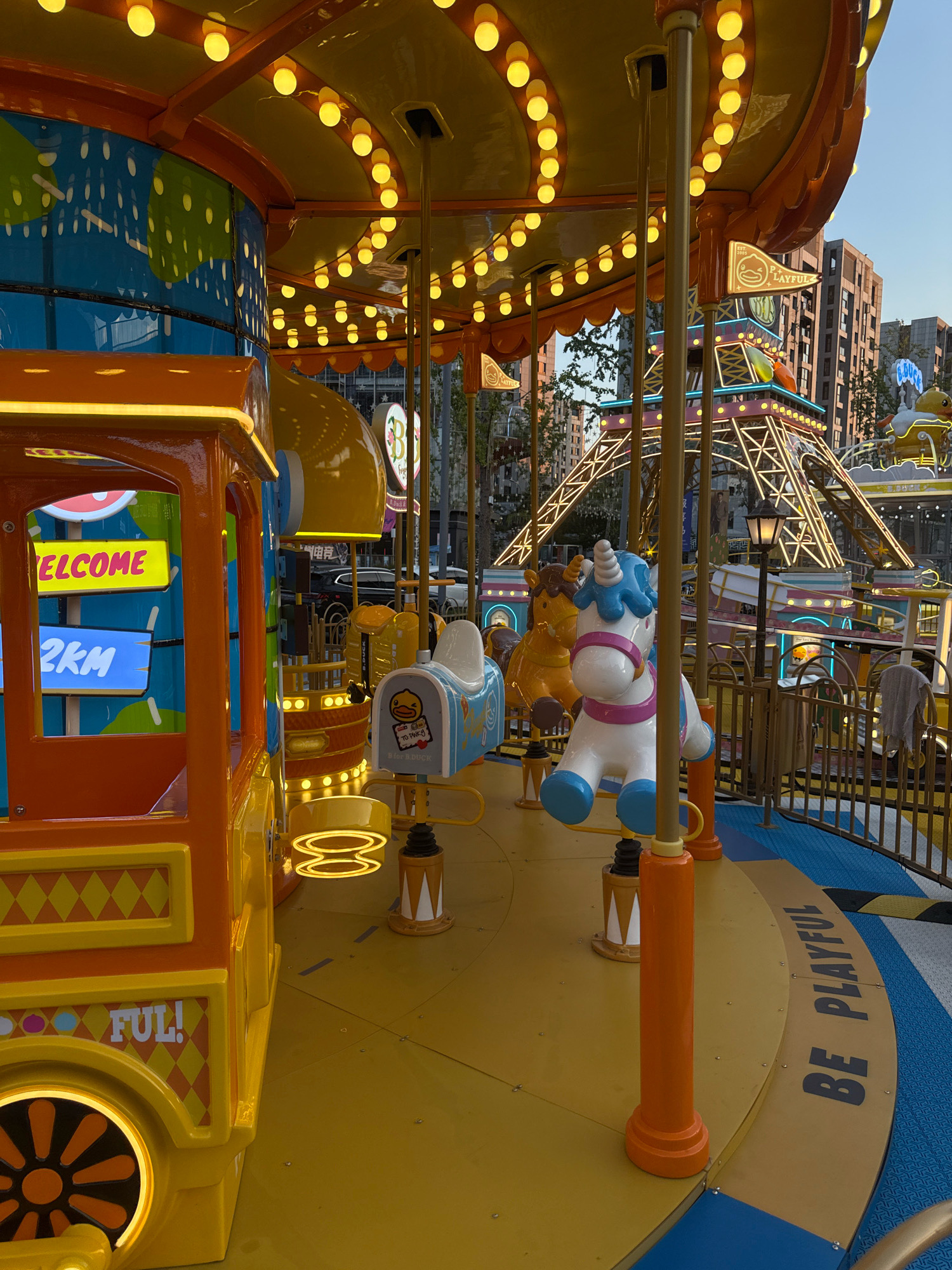 The image shows an outdoor amusement park, likely a carousel, in the city of Yiwu, China. The carousel features brightly colored vehicles and carousel animals, including a unicorn, horses, and a duck-themed ride. The structure is adorned with numerous yellow lights. In the background, the Eiffel Tower replica and other amusement park attractions are visible, along with modern buildings. The ground is a mix of yellow and blue patterned surfaces. A sign on the edge of the carousel reads "BE PLAYFUL". The scene is well-lit, suggesting it is either daytime or the park is illuminated for evening operation. There are no people clearly visible in the image.
