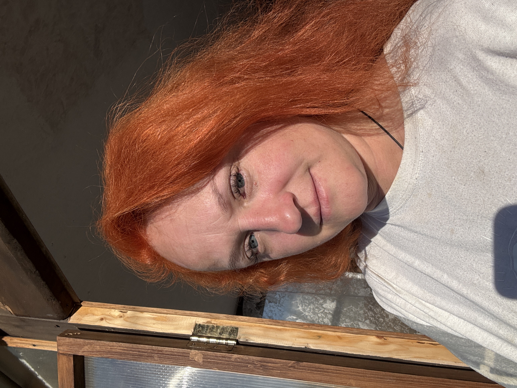 A woman with long, red hair and blue eyes is looking into the camera. She is wearing a white t-shirt. Her face is illuminated by sunlight. The background shows a wooden frame and a translucent panel, suggesting a window or door. The location is unknown, and there is no information in the image that indicates it is Chaikovsky, Russia.