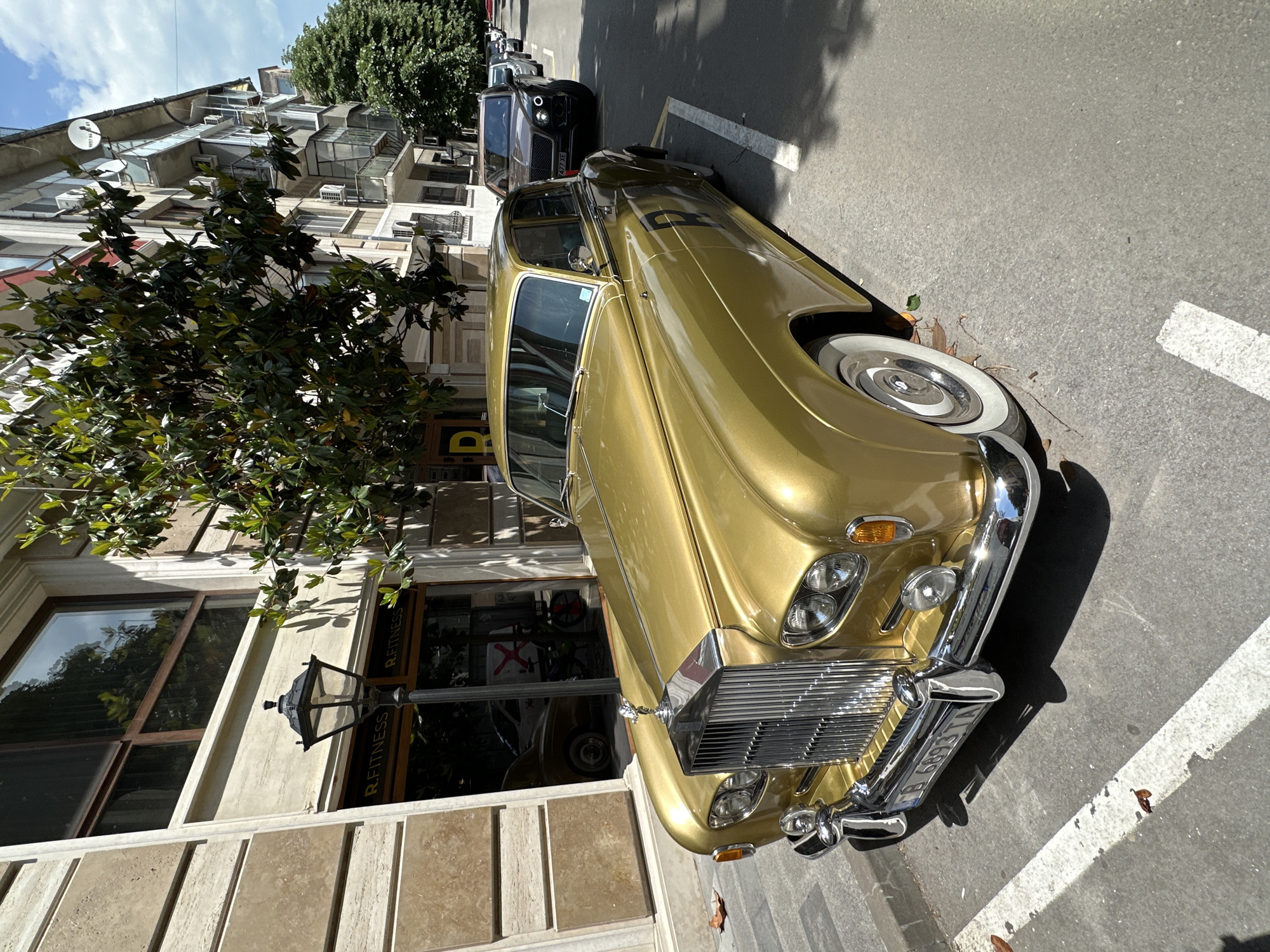 A slightly rotated outdoor shot shows a vintage gold Rolls Royce parked on a street in Varna, Bulgaria. The car faces the right of the frame and is parked along a curb lined with white parking lines. A black Bentley is parked behind the Rolls Royce. Buildings line the street. To the left, part of a building's facade is visible, including a large window reflecting the street scene and a sign that reads "R. FITNESS." A tree with dense green foliage grows near the building. The sky is visible, appearing bright and sunny. The overall scene suggests a well-maintained urban environment.