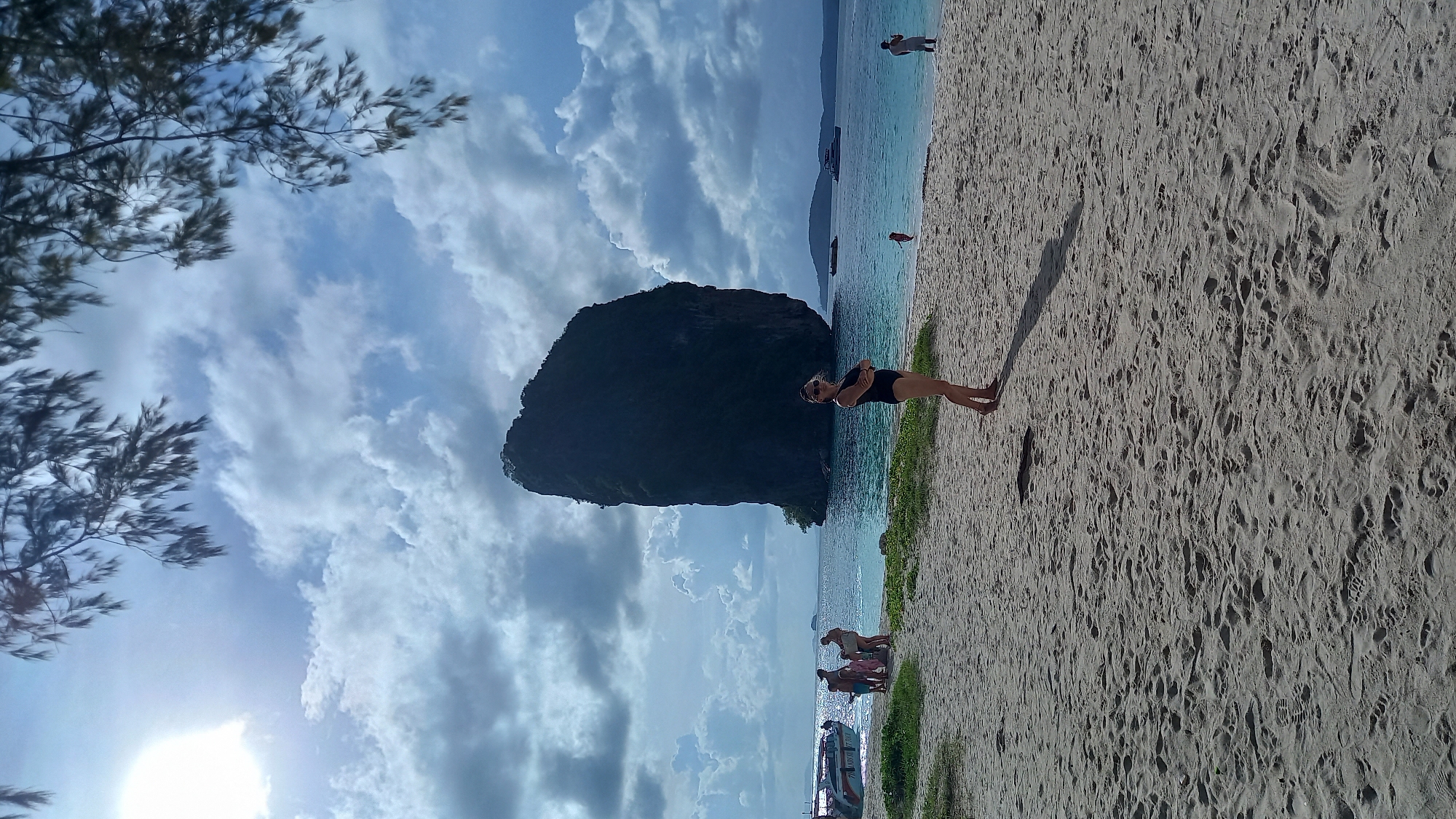 The image shows a beach scene in Ban Ao Nang, Thailand. A woman in a black swimsuit stands on the sandy beach near the water's edge. A large limestone rock formation rises out of the water behind her. In the background, other people are visible on the beach and in the water. A few boats are also seen in the water. A tree branch is on the left side of the picture. The sky is cloudy, with some blue visible. The sun is shining in the bottom left corner of the picture.