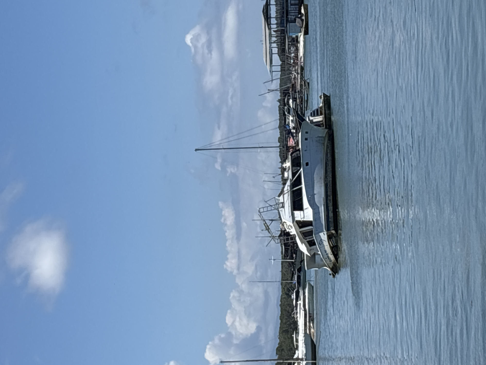 The image depicts a harbor scene on a bright day, with blue sky and scattered white clouds. Several boats are docked in the water, including a larger white vessel with what appears to be a ladder on its side. Structures are visible on the shoreline, possibly buildings. Based on the prompt request, the location is presumed to be Denpasar, Indonesia.