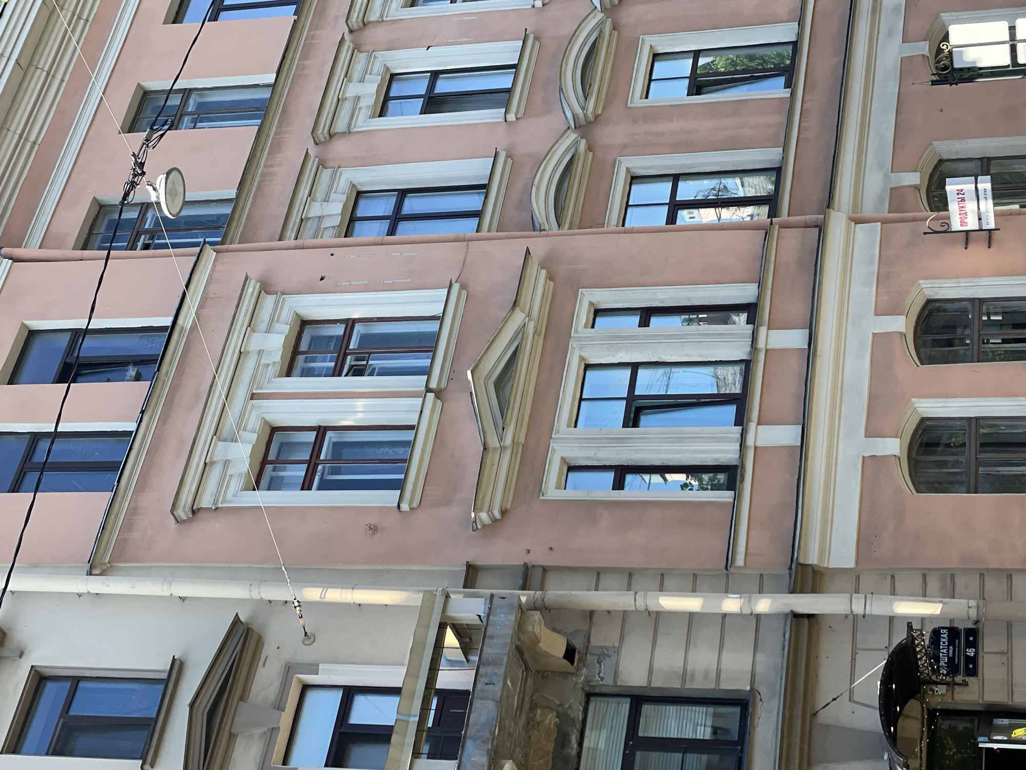 The image shows a section of a light pink and beige building with multiple windows and architectural details. A street lamp and associated wiring are visible on the facade. A street sign, labeled "Kryushatskaya" in Russian and "46," is attached to the building near its base. The building's architectural style and signage suggest that it is located in Saint Petersburg, Russia.