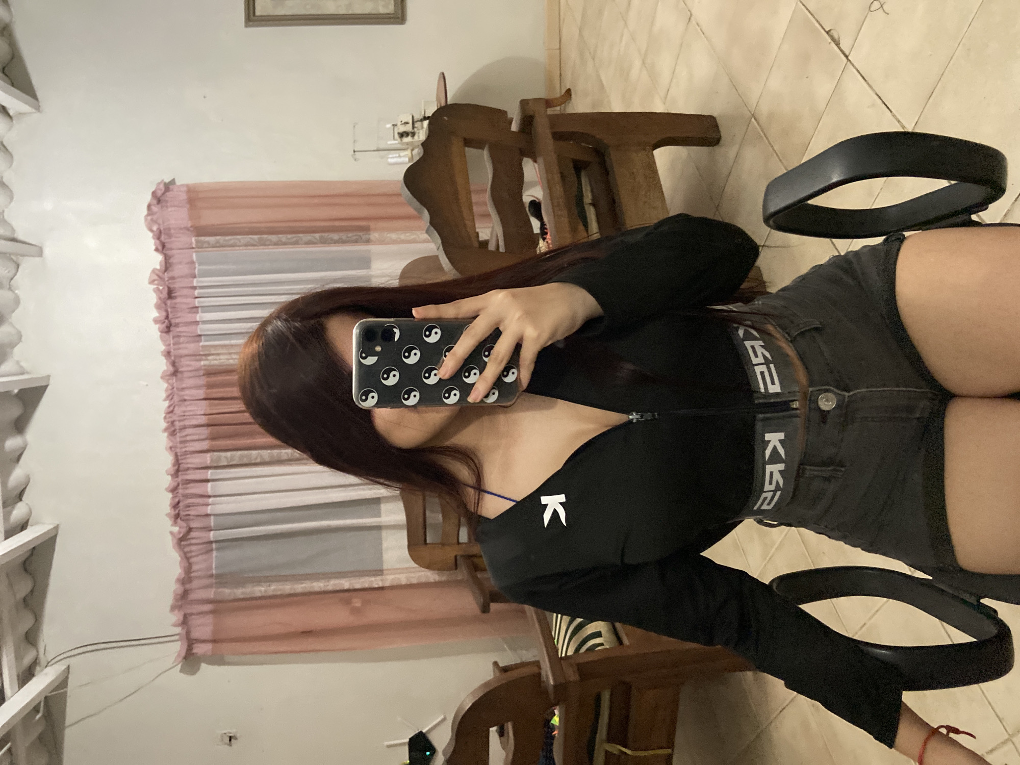 A person is taking a selfie in what appears to be a bedroom or living room. The person is holding a smartphone with a case that has yin-yang symbols printed on it. The person is wearing a black long-sleeved top with a white "K" logo on the chest and dark-colored denim shorts with a belt. A black belt lies near the person's waist on the right side of the frame. The background includes wooden furniture, a window with pink curtains, and a tiled floor. The location is indoors, but the city of Juan Griego, Venezuela, is not discernible from the image.