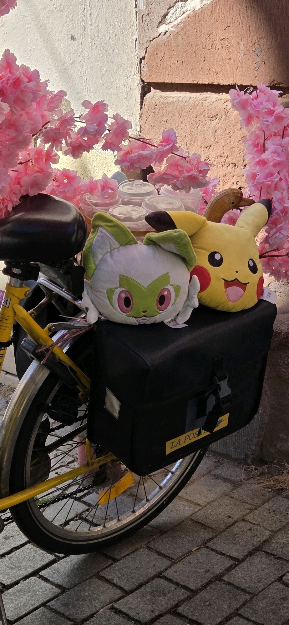 In Strasbourg, France, a yellow bicycle is parked outdoors. The bicycle has a black bicycle seat and a black pannier bag mounted on the rear rack. On top of the pannier bag are two stuffed toys: one resembles Pikachu, a popular character from the Pokémon franchise, and the other is a green and white character. Behind the bicycle, several translucent cups with lids are visible, suggesting they may contain beverages. The scene is adorned with artificial pink cherry blossoms, creating a decorative backdrop. The bicycle and its contents are positioned against a textured wall made of both light-colored plaster and reddish-orange bricks. The ground is paved with rectangular stone blocks. The overall impression is a charming and slightly whimsical display, possibly set up for an event or a personal decorative arrangement.