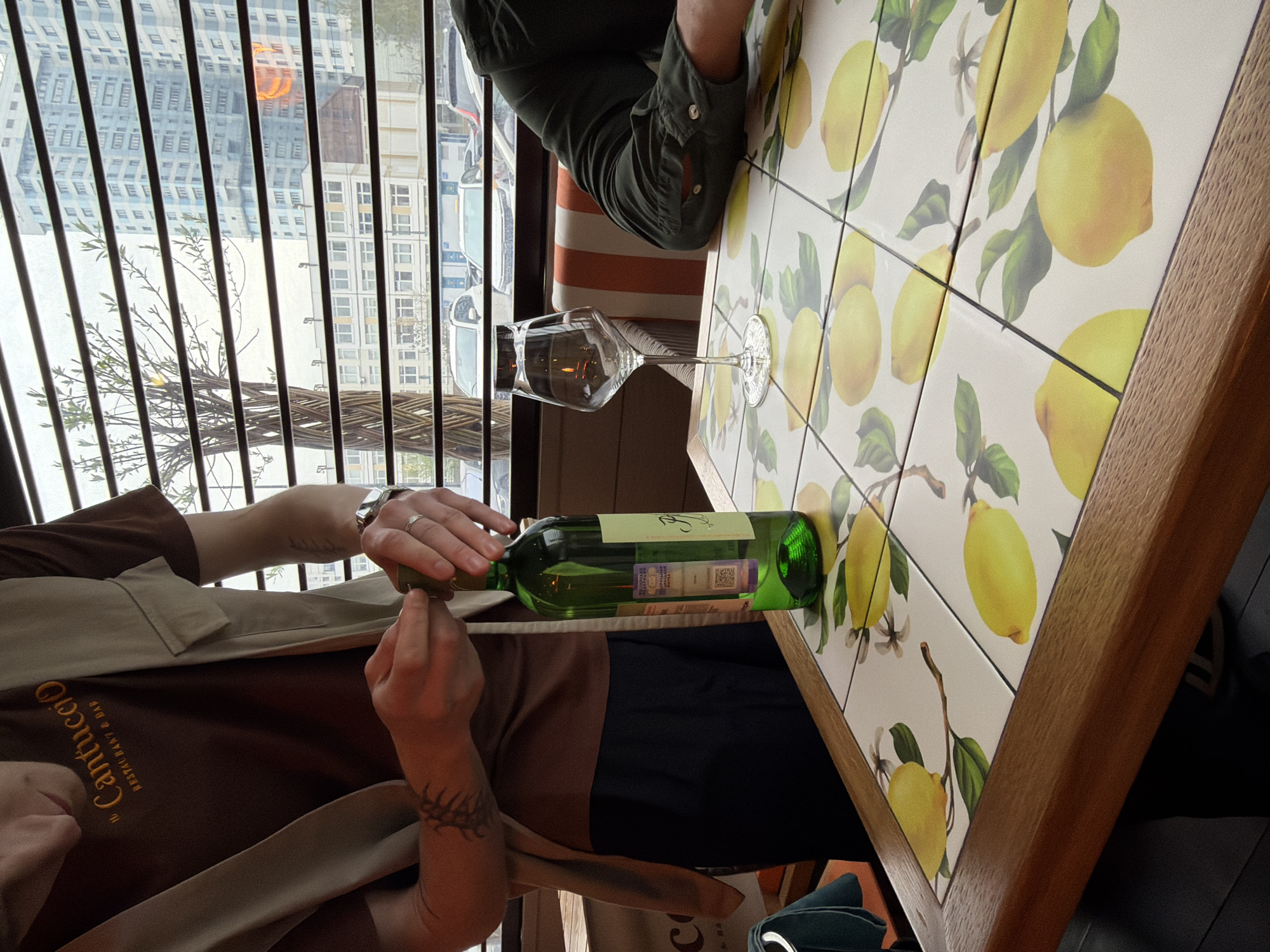 The image depicts a scene at a restaurant, likely in the city of Khimki, Russia, indicated by the "Cantuccio Restaraunt & Bar" inscription on a person's shirt. The setting is indoors, with a view of buildings visible through a window. At least two people are present. One person is wearing a brown and beige shirt, with a visible tattoo on their arm, and is seemingly opening a bottle of wine. Another person, wearing a green shirt, is seated across the table. A wine glass sits on the table, and the table itself is decorated with a lemon pattern. The bottle of wine is green and appears to be sealed.