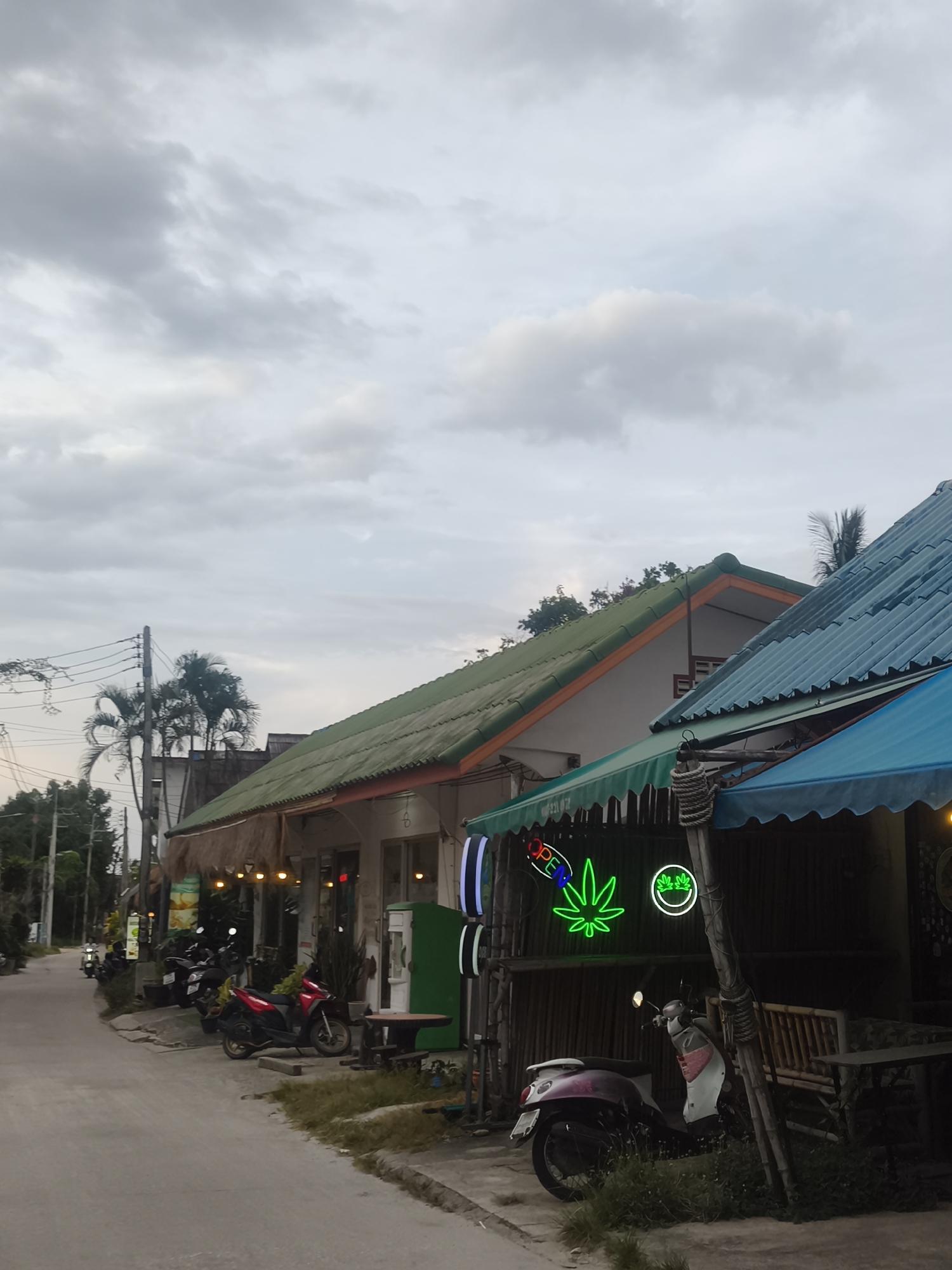 The image depicts a street scene in a tropical location, likely Ko Samui District, Thailand, given the architecture and vegetation. The sky is overcast with gray clouds. On the right side of the street, there are several buildings with corrugated metal roofs, one a light blue and another green. One building has a thatched awning over its entrance, and neon signs, including a green cannabis leaf and an "OPEN" sign. In front of the buildings, several motorcycles are parked. The street itself is paved with asphalt and appears to be a local road. Palm trees and other lush vegetation are visible on the left side of the image, along with utility poles and wires. The overall impression is of a commercial street in a relaxed, possibly tourist-oriented area, in the evening or on a cloudy day.