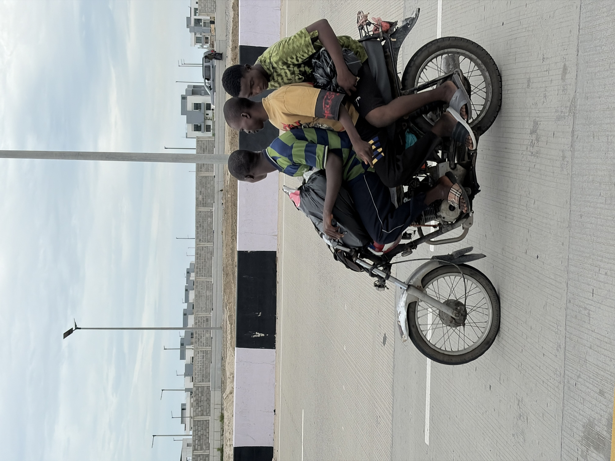 Three young men are riding on a motorcycle in Igboefon, Nigeria. The man in the front is driving, wearing a green and blue striped shirt and black pants. The middle passenger is wearing a yellow t-shirt with red lettering and black shorts. The passenger in the back is wearing a green camouflage t-shirt and black pants. All three are wearing sandals. They are carrying black plastic bags on the motorcycle. The motorcycle is traveling on a paved road with white lines. In the background, there are buildings under construction, a grey brick wall, and streetlights. The sky is overcast.
