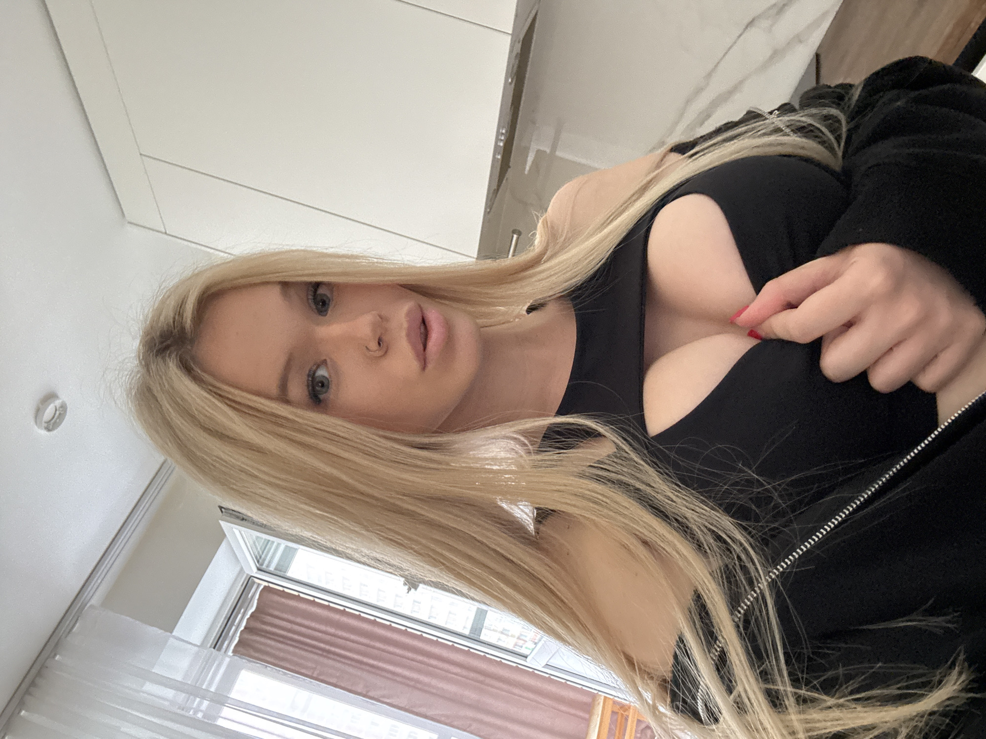 A young woman with long blonde hair and blue eyes, wearing a black cutout top and a black zip-up jacket, is taking a selfie indoors. Her left hand is adjusting her top. The background shows a window with white curtains and a pink curtain, as well as kitchen cabinets and a countertop. The image appears to have been taken in an apartment in Saint Petersburg, Russia.