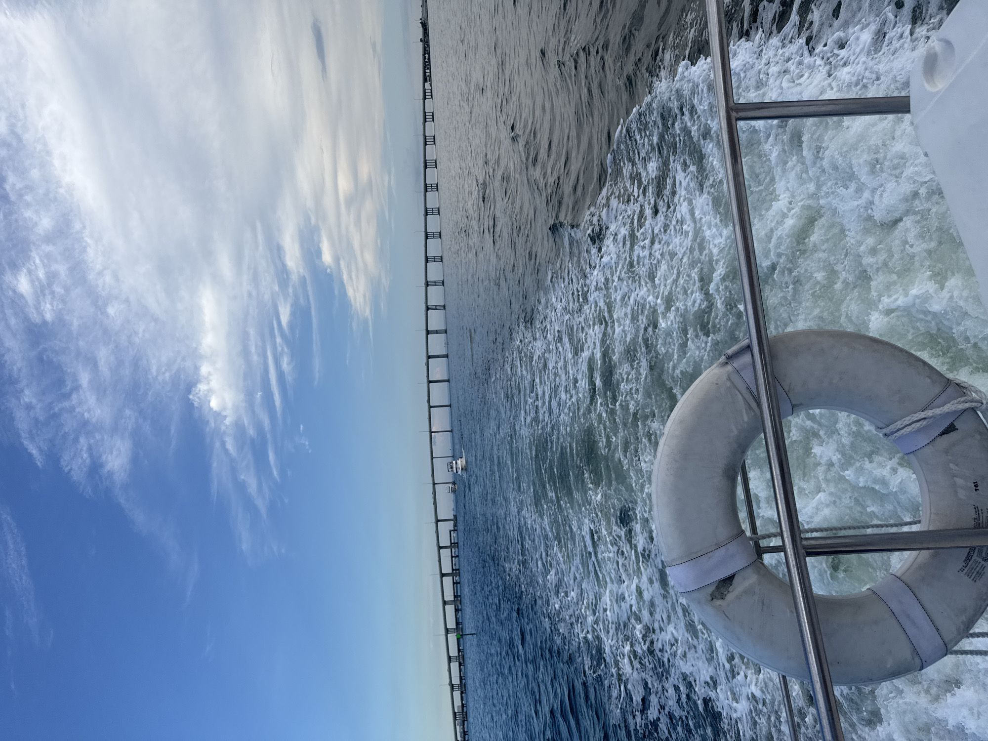 A boat is moving away from the camera, leaving a wake of white foamy water. In the foreground, a life preserver is visible, mounted on a metal railing. In the background, a long, multi-span bridge stretches across a wide body of water. The sky is blue with scattered clouds. The scene appears to be on a bay or ocean, likely near a city. While the image captures elements of a maritime environment and infrastructure common to coastal cities, there are no specific visual cues to definitively place it within Miami, United States of America.