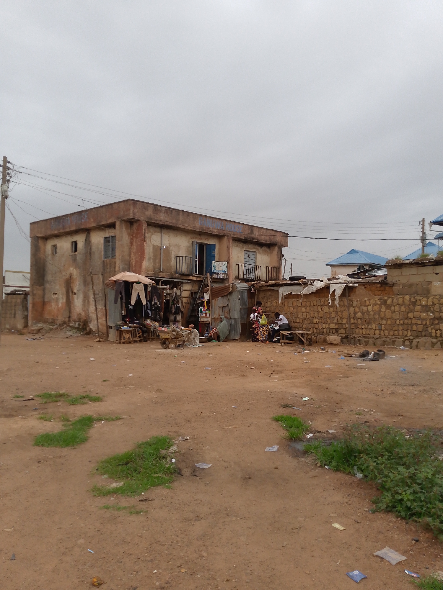 The image depicts a street scene in Bukuru, Nigeria, under an overcast sky. In the background, a two-story, weathered building stands adjacent to a market-like area. Clothing items hang from an improvised shelter in front of the building. People are gathered nearby; some are seated and others are interacting. A stone wall runs along the side, next to a blue-roofed house. The foreground consists of an unpaved, sandy area with scattered debris and patches of vegetation. A utility pole with wires is visible.