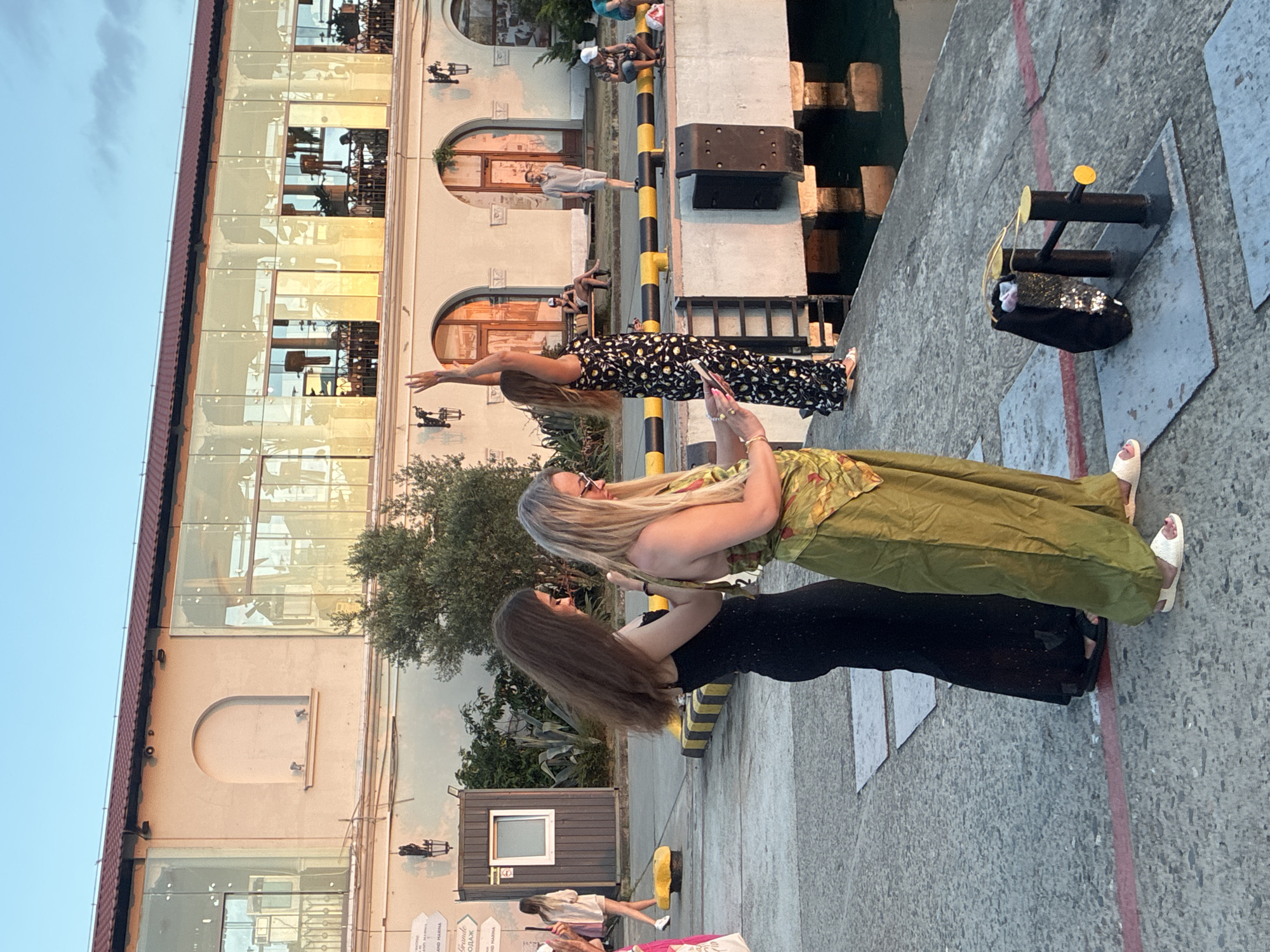 The image depicts a waterfront scene in Sochi, Russia, likely near the port or a promenade. In the foreground, three women are visible. Two women are standing close together, facing away from the camera, their backs to the viewer. The woman on the left is wearing a long, black, sleeveless dress. The woman on the right is wearing a patterned top with wide-leg olive-green pants. She appears to be looking down at a phone or similar device. To the left of these two women, a third woman, also facing away from the camera, is wearing a light-colored dress and white sneakers, and is walking away.
Behind the women, a building with a modern design is visible, featuring large glass windows that reflect the warm, golden light of what appears to be late afternoon or early evening. The building has arched entrances and an ornate facade.
In the middle ground, several people are seated on benches or standing, further away from the camera. A man in a light-colored shirt and shorts stands near an arched doorway. Further to the right, two people are seated on a bench.
The foreground area where the women are standing is a concrete pier or walkway, bordered by a yellow and black striped barrier. In the lower right portion of the image, a black mooring bollard with yellow caps is present, with a black bag resting on it. The water is visible on the right side of the image, reflecting the building's exterior. The overall lighting suggests a pleasant, warm day in Sochi.