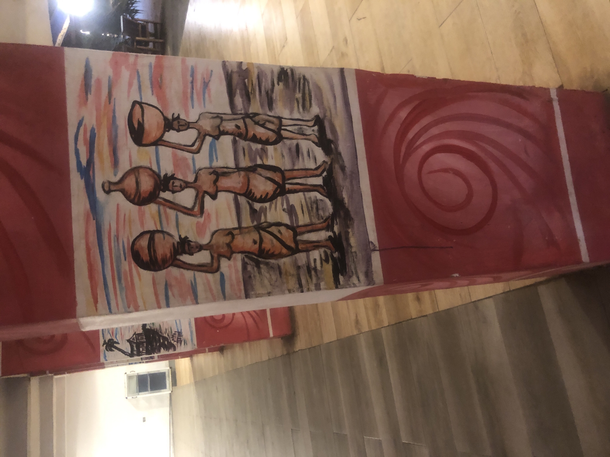 A close-up shot captures a painted pillar with an artistic depiction of three women carrying water vessels on their heads. The pillar is decorated with red as the primary color, adorned with white stripes and a swirling pattern at the bottom. The painting itself features a colorful sky in shades of pink, orange, blue, and white, contrasting with the earthy tones of the ground. The women are rendered in warm, skin-like colors, dressed in simple wraps, carrying spherical and bowl-shaped vessels. To the left of the pillar, a glimpse of a building with white walls and a glass door is visible. The floor is a combination of light brown and grey wooden planks. The scene suggests an indoor setting, possibly a hallway or an entrance area, with the backdrop implying an outdoor environment or a stylized representation of one. The painting is done in a simplistic, illustrative style. The location is specified as Iwesolu, Nigeria.