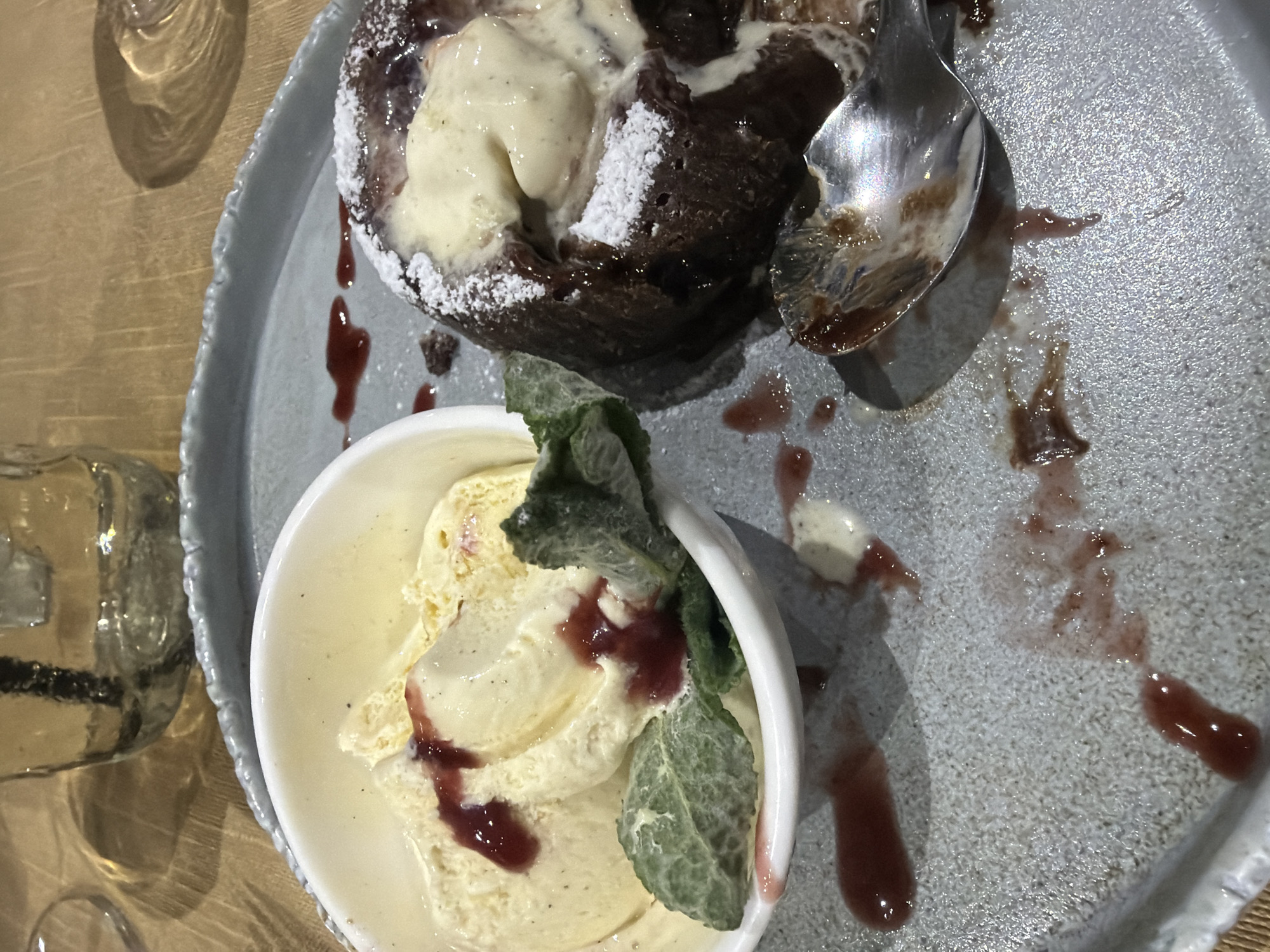 A top-down, close-up photograph displays a dessert on a light grey, textured ceramic plate with an irregular rim, resting on a light-colored wooden table. The central subject is a dark chocolate dessert, likely a lava cake, generously topped with a scoop of melting vanilla ice cream and dusted with white powdered sugar. A silver spoon is partially inserted into the chocolate dessert, indicating it is being or has recently been consumed.
Adjacent to the chocolate dessert, a small white bowl holds additional scoops of vanilla ice cream. This ice cream is garnished with a reddish-purple berry compote or sauce and a fresh green mint sprig. Streaks and drips of this berry sauce, along with melted ice cream, are visible on the grey plate, suggesting the dessert is in the process of being enjoyed.
In the upper left portion of the image, the reflections and outline of a clear glass object, possibly a drinking glass, are visible on the wooden table surface. The scene is brightly lit, typical of an indoor dining setting, possibly a restaurant or cafe. No visible text is present in the image.