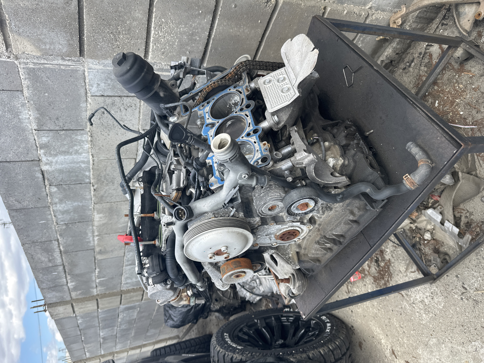 The image shows a partially disassembled car engine mounted on a black metal stand. The engine is missing its cylinder head and shows the head gasket. The engine appears to be in a used condition with some rust. The location appears to be outdoors. The background includes a cinder block wall, spare tires, and general debris. The image was likely taken in Piperevo, Bulgaria.