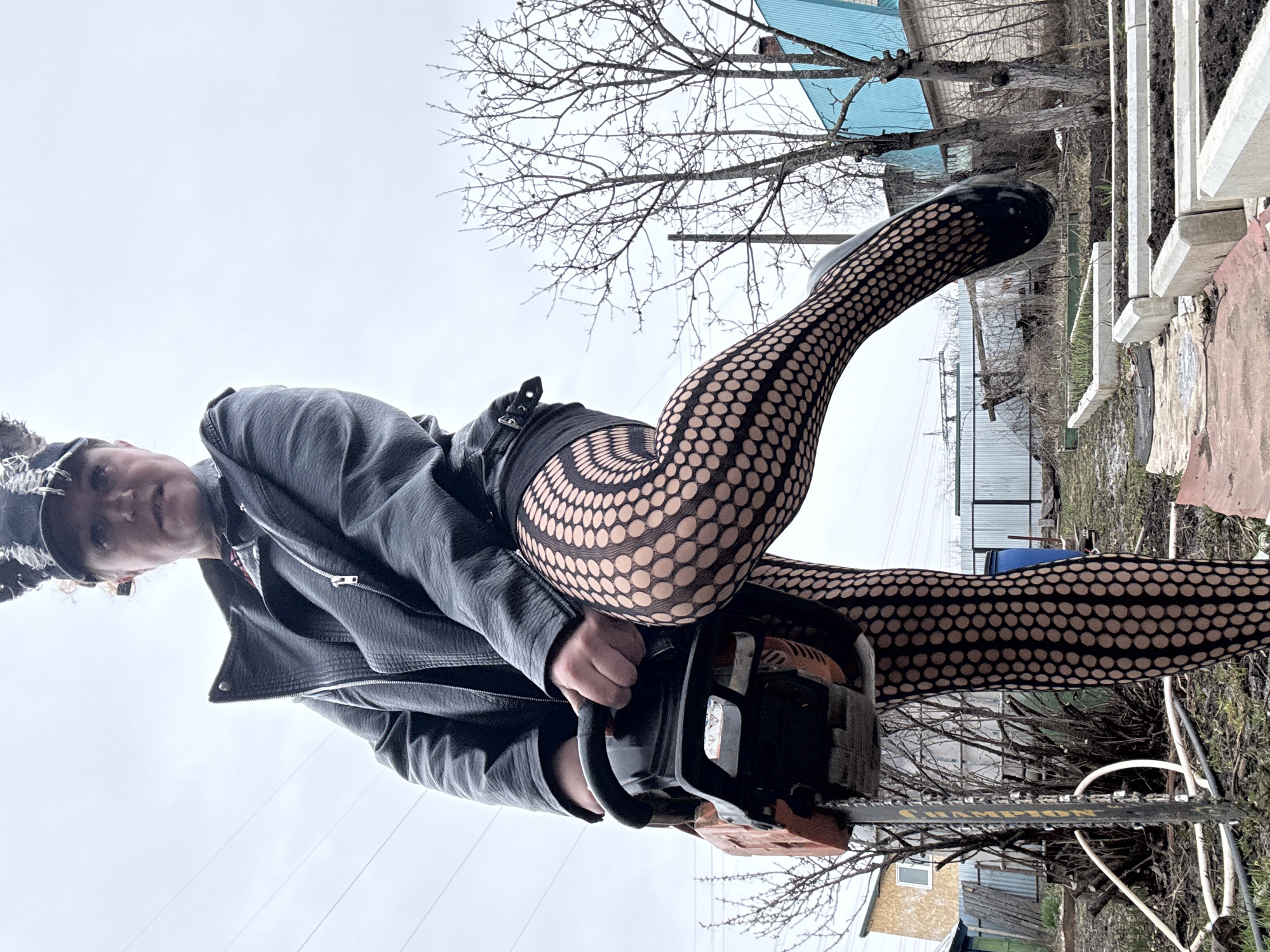 A person wearing a black leather jacket, black shorts, and patterned fishnet stockings is holding an orange and black chainsaw in a yard. The background includes a blue roofed building, bare trees, a metal fence, and other residential structures. The location is Chaikovsky, Russia.