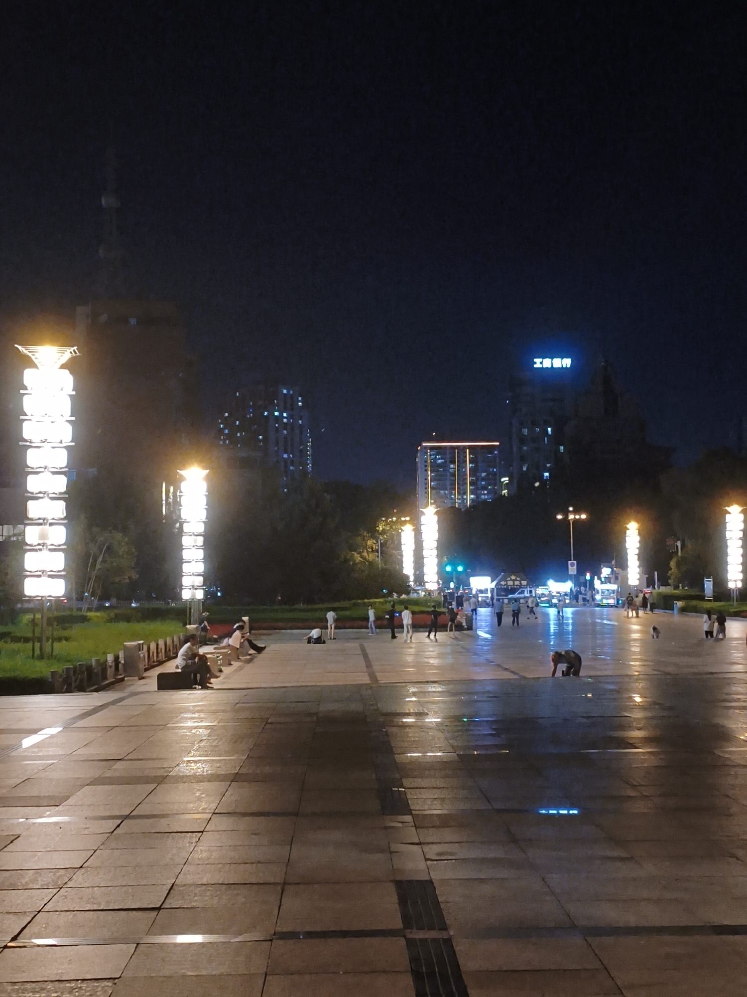 The image captures an urban plaza scene at night in Xi'an, China, specifically the area in front of the Xi'an Museum. Tall, illuminated light poles line the plaza, casting a bright glow on the wet, tiled ground. Several people are present, some sitting on benches, others walking or standing. In the background, illuminated buildings and a distant tower are visible against the dark night sky. One building features bright blue neon signage that reads "工商银行", which translates to "Industrial and Commercial Bank of China". The wet pavement reflects the lights, creating a glossy, somewhat blurred effect. A person is crouched low in the foreground.
