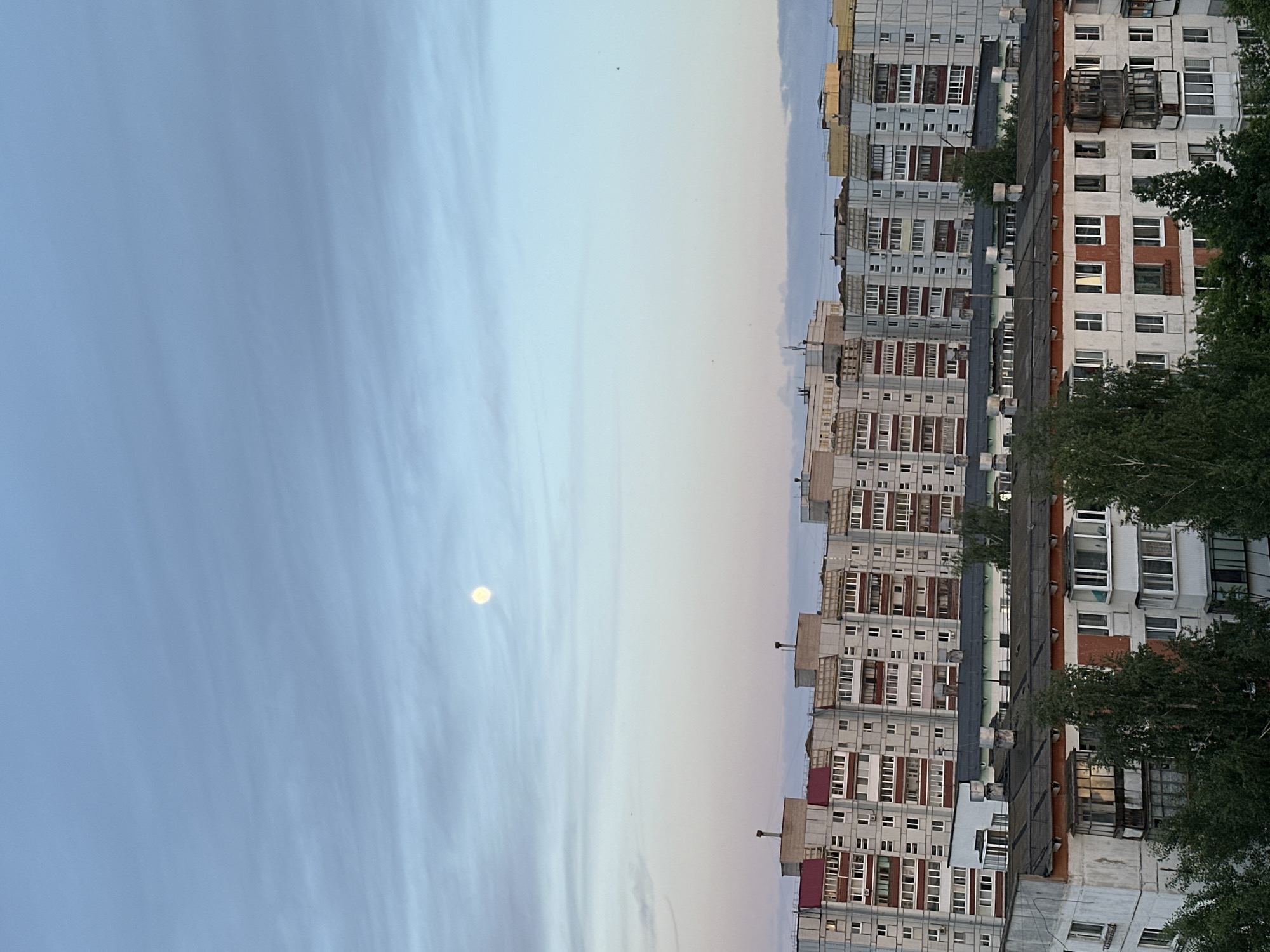The image is an urban scene in what appears to be the city of Chaikovsky, Russia. It's a view from a high vantage point, likely an upper story of a building. The focus is on a row of Soviet-era apartment buildings, characterized by their repetitive window patterns, drab colors, and flat roofs. Some buildings have balconies, and a few show signs of disrepair or aging. The buildings are situated in a densely populated area, evidenced by the close proximity of the structures and the presence of utility infrastructure on the rooftops. Trees are visible near the buildings, adding some greenery to the scene. The sky takes up about half of the image and it is a mix of blue and grey. There is also a single distant orb in the sky, and what appears to be birds flying in the distance. The overall mood is one of a calm, overcast day.