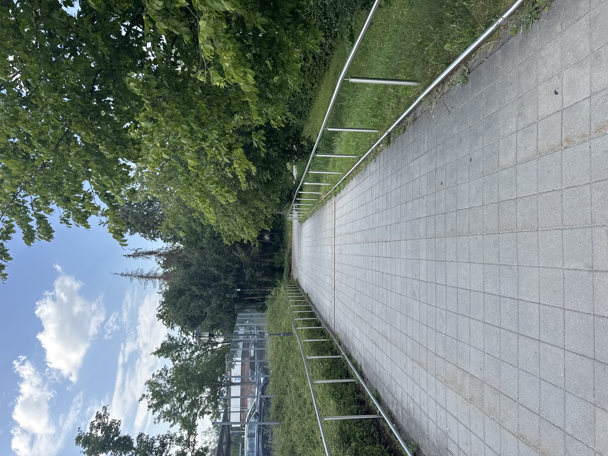 The image depicts a paved walkway curving to the right, bordered by a metal railing on the left. Lush green trees and foliage flank the walkway, with a patch of grass and a chain-link fence visible beyond the railing. In the distance, parked cars are visible behind the fence, suggesting an urban or suburban setting. The sky above is a bright blue, dotted with white clouds, indicating fair weather during the daytime. The scene is a peaceful outdoor environment, possibly a park or a path alongside a public area. No people are visible in the image, and there is no visible text. The image appears to be taken in Höhenkirchen-Siegertsbrunn, Germany.