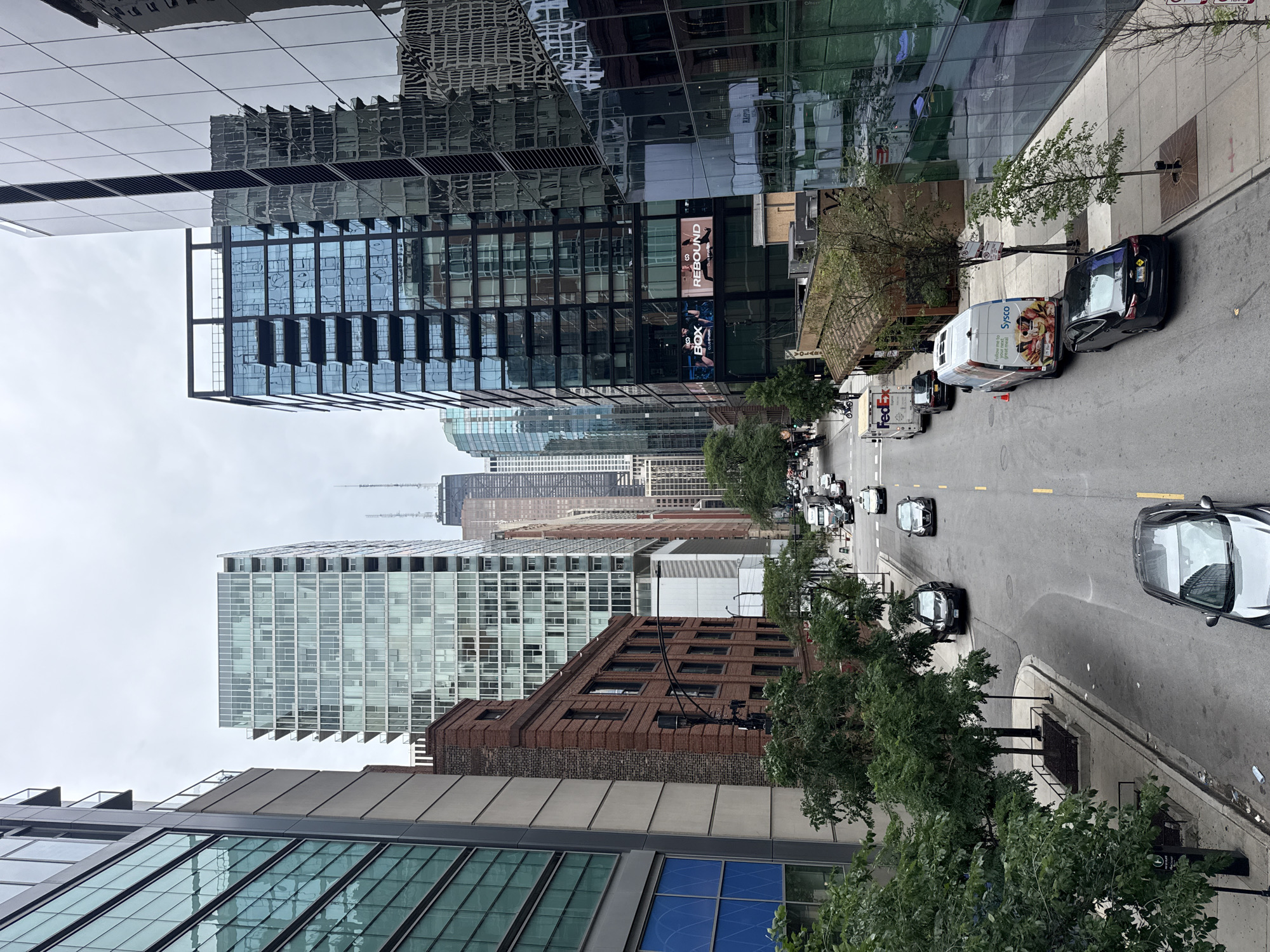 This image depicts a city street scene, likely in Chicago, USA, with a focus on urban architecture and vehicular traffic. The perspective is from a high vantage point, looking down at the street and surrounding buildings.
The primary subjects are the modern skyscrapers with glass facades, reflecting the overcast sky and the buildings opposite. One building features prominent signage for "REBOUND" and "BOX CLUB". The street is populated by several cars, including a black sedan, a white van with "Sysco" branding and food imagery, and a FedEx truck. A few pedestrians are visible on the sidewalk, though their details are indistinct. The activity is the normal flow of city life with vehicles moving along the road and people present on the sidewalks.
Notable details include the overcast weather, suggested by the muted light and the appearance of the sky. The time of day appears to be daytime. The presence of tall, contemporary buildings with extensive glasswork strongly suggests a major metropolitan area, consistent with the provided location context. The overall mood is neutral, documenting an urban environment.
Visible text includes "REBOUND", "BOX CLUB", "Sysco", and "FedEx".