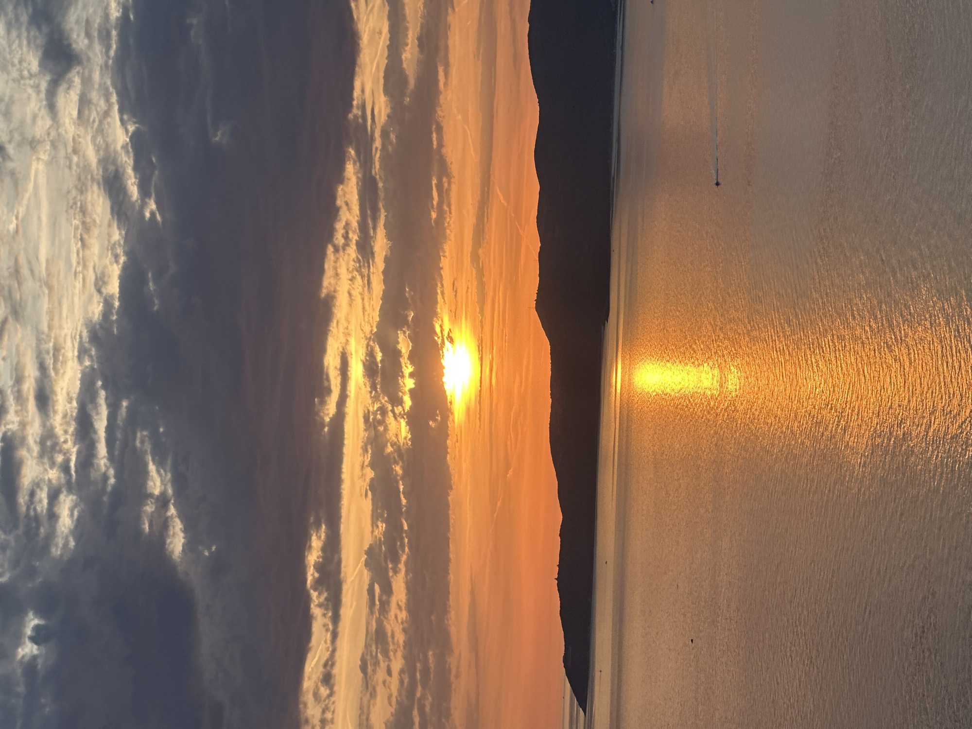 The image displays a sunset scene over a body of water identified as being in Rijeka Reževići, Montenegro. The sun, a brilliant yellow-orange sphere, is situated slightly left of the vertical center, resting just above a dark, continuous landmass. The sky above is characterized by an array of clouds: the upper portion features dense, dark grey cumuliform clouds with lighter, warm-toned patches visible through gaps, while the middle and lower sky glow with intense orange and yellow hues around the sun. Several faint, linear contrails are visible against the colorful sky.
The landmass, appearing as a dark silhouette of rolling hills or mountains, extends horizontally across the mid-ground. A small, distinct structure, possibly a lighthouse or building, is visible atop a peak on the left side of this landmass.
The lower half of the image is dominated by the water, which reflects the sun's intense light, creating a shimmering, golden-orange path directly beneath the sun and extending towards the foreground. The water's surface exhibits subtle ripples and texture. In the mid-lower right area of the water, a small, dark boat with a visible wake is moving towards the right. Another minuscule, dark object is visible on the water further to the left in the distance, along with several other indistinct dark specks. No human figures are present.