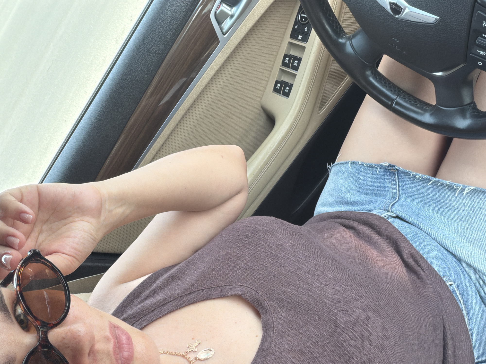 A selfie-style, full shot of a fair-skinned woman inside a Genesis car. She wears a maroon top and denim shorts, resting her left arm near her face, sunglasses perched on her head. A gold necklace is visible on her chest. The car interior features beige leather and wood trim. The steering wheel bears the Genesis logo. The location in Chaikovsky, Russia, cannot be determined from the image.