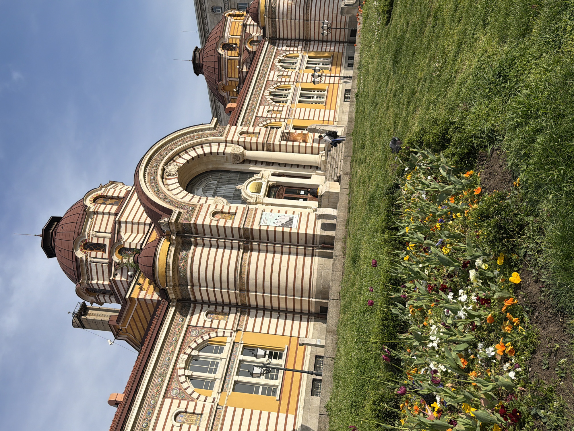 An outdoor, brightly lit, daytime shot captures an ornate, historic building with a large green lawn and flowerbed in the foreground. The building, likely a public structure like a museum or bathhouse, features distinctive striped facades in alternating light and dark bands, primarily yellow, orange, and reddish-brown. It is adorned with multiple domed roofs, grand arched windows, and intricate decorative patterns around the arches and cornices, suggesting a Neo-Byzantine or Moorish Revival architectural style.
In the mid-ground, a person is seated on the stone steps leading to the building's main entrance. On the right side of the image, a vibrant flowerbed bursts with colorful blooms, including yellow, orange, white, and purple flowers, some resembling tulips. Beyond the flowerbed, a lush green lawn extends. A pigeon is visible on the grass near the flowers. The sky is clear blue with sparse clouds, indicating pleasant weather. A poster with images of two figures, possibly for an exhibition or performance, is affixed to the building's wall near the entrance, though the text is not clearly legible. The scene conveys a tranquil atmosphere in a grand urban setting.