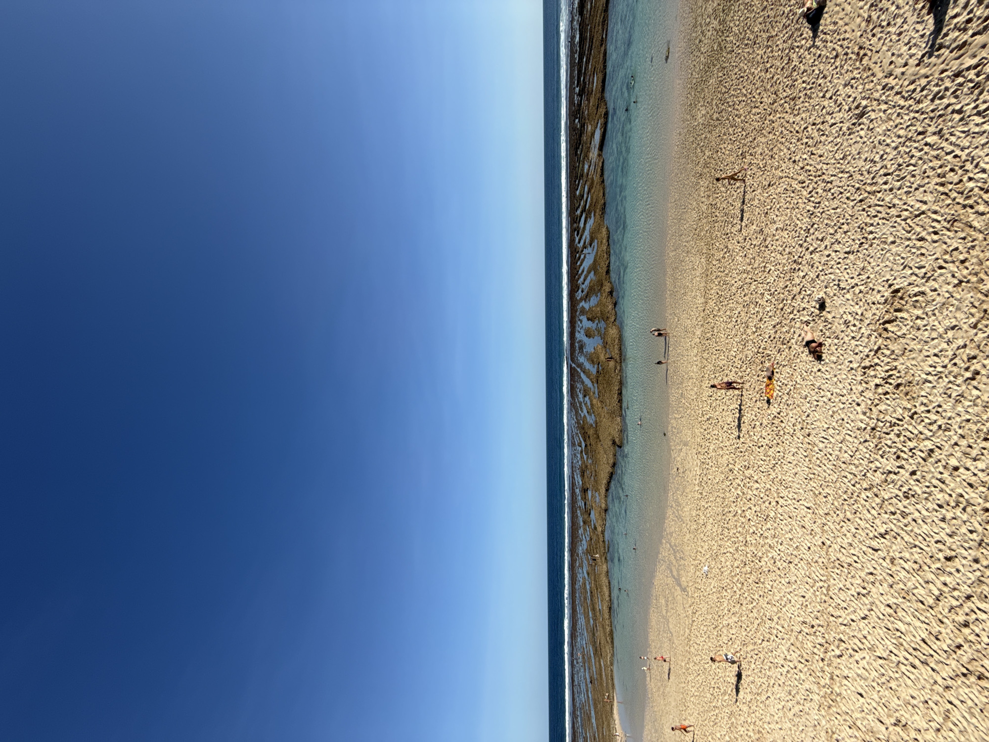 This media file captures a picturesque, expansive view of a tropical beach under a clear, bright sky. The image is oriented vertically but depicts a horizontal landscape.
The upper portion of the frame is dominated by a vast, cloudless blue sky, transitioning from a deeper blue at the top to a lighter hue towards the horizon. A distinct, straight line marks the horizon, separating the sky from the ocean.
Below the sky, a strip of deep blue ocean meets a wide, shallow area of incredibly clear, turquoise water. This clear water washes over extensive, dark brown rock formations (likely coral or volcanic rock) that are exposed along the shoreline, suggesting a low tide. These textured formations create natural pools and channels of water.
The lower half of the image features a wide, sandy beach with golden-brown sand, showing ripples and patterns created by the tide and wind. Several small figures of people are scattered across the beach and in the shallow water. Some individuals are seen wading or swimming in the clear water near the rock formations, while others are relaxing or walking on the sand. Long, distinct shadows cast by the people indicate strong, overhead sunlight, suggesting it is likely late morning or early afternoon.
The overall scene conveys a peaceful, leisurely atmosphere on a beautiful, sunny day at a tropical beach. No visible text is present in the image. The setting is consistent with a vibrant coastal area such as Kuta Selatan, Indonesia.