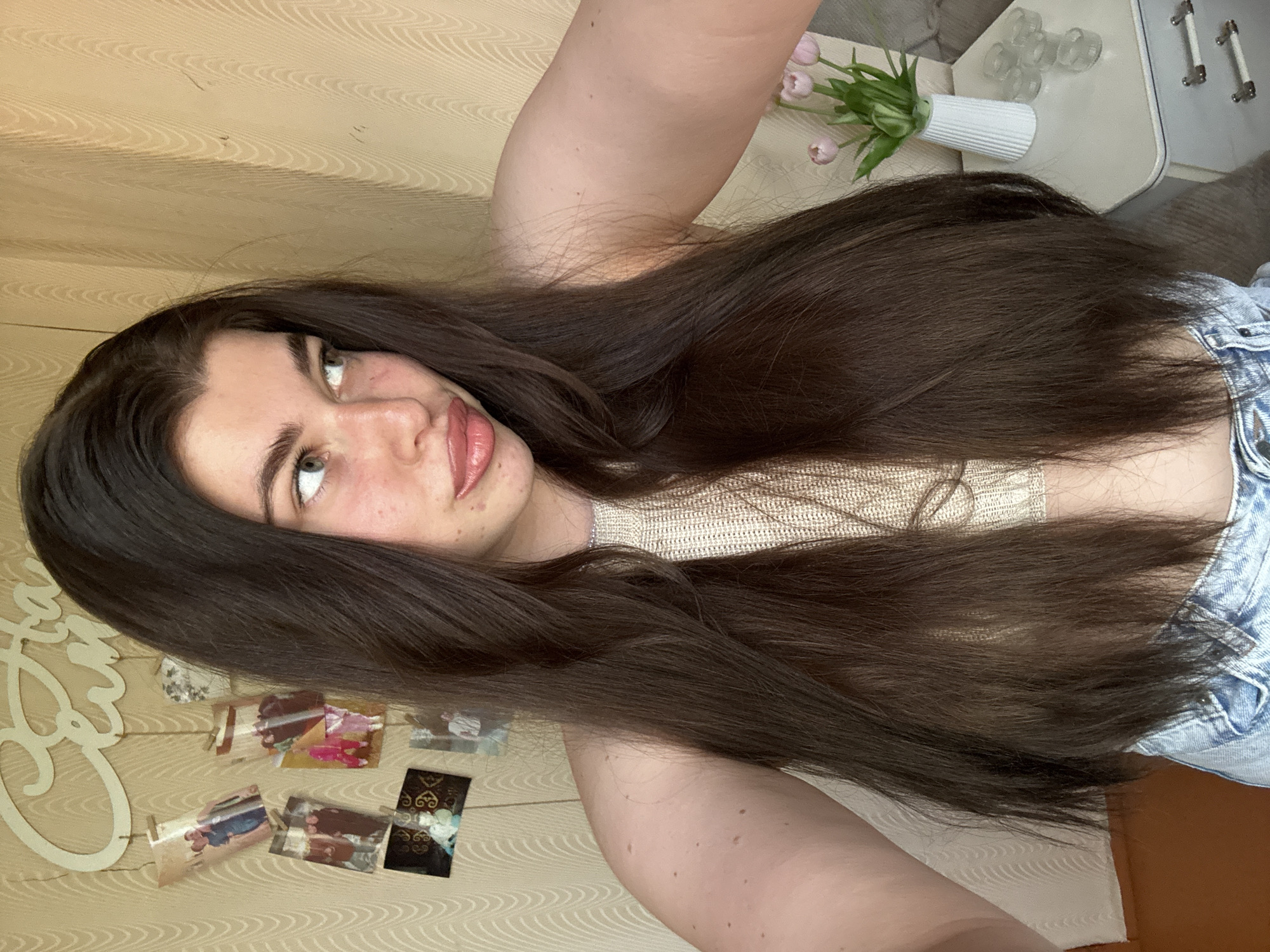 A young woman with long, dark brown hair is taking a selfie. She is wearing a beige top and denim shorts. In the background, there is a wall decorated with photos and a cursive word art. There is a side table with a vase of tulips and some glasses. The image does not provide enough information to determine the location in the city of Мещёры, Russia.