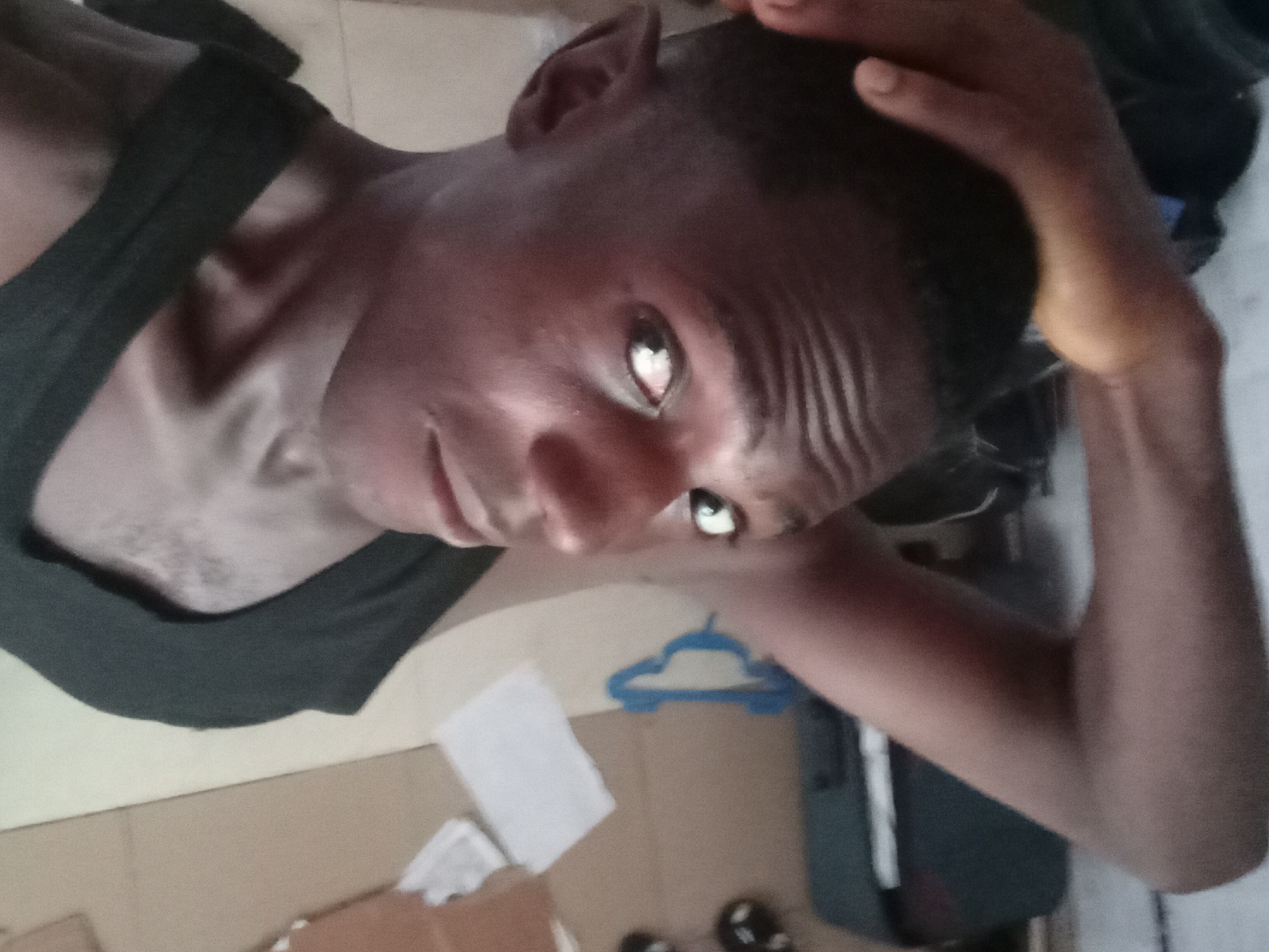 A low-angle, close-up shot of a person with dark skin wearing a black tank top. The person is looking directly at the camera with their right hand on the top of their head. The background includes a white piece of paper, a blue clothes hanger, shoes, and part of a suitcase on a light-colored tiled floor. The location is Akwete, Nigeria.