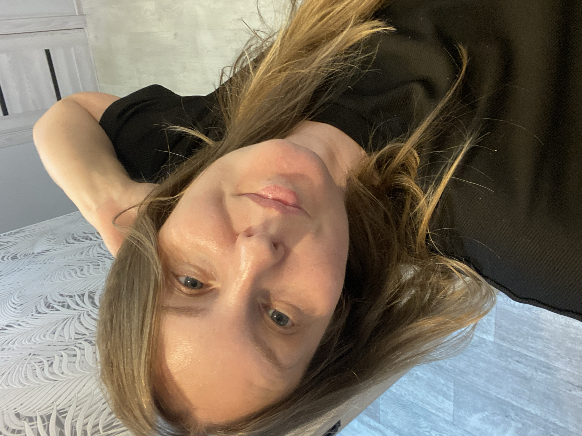The image shows a close-up, selfie-style photograph of a woman from the chest up. She has light brown hair that falls past her shoulders and is wearing a black t-shirt. Her left arm is bent behind her head, with her hand resting on her neck. She has fair skin, blue eyes, and appears to be smiling slightly. The background is out of focus, but appears to be an indoor setting with patterned wallpaper on the left and what looks like a door or closet on the right. The floor has a light, wood-grain pattern. There is no identifiable information about the specific location within the city of Noviy, Russia.