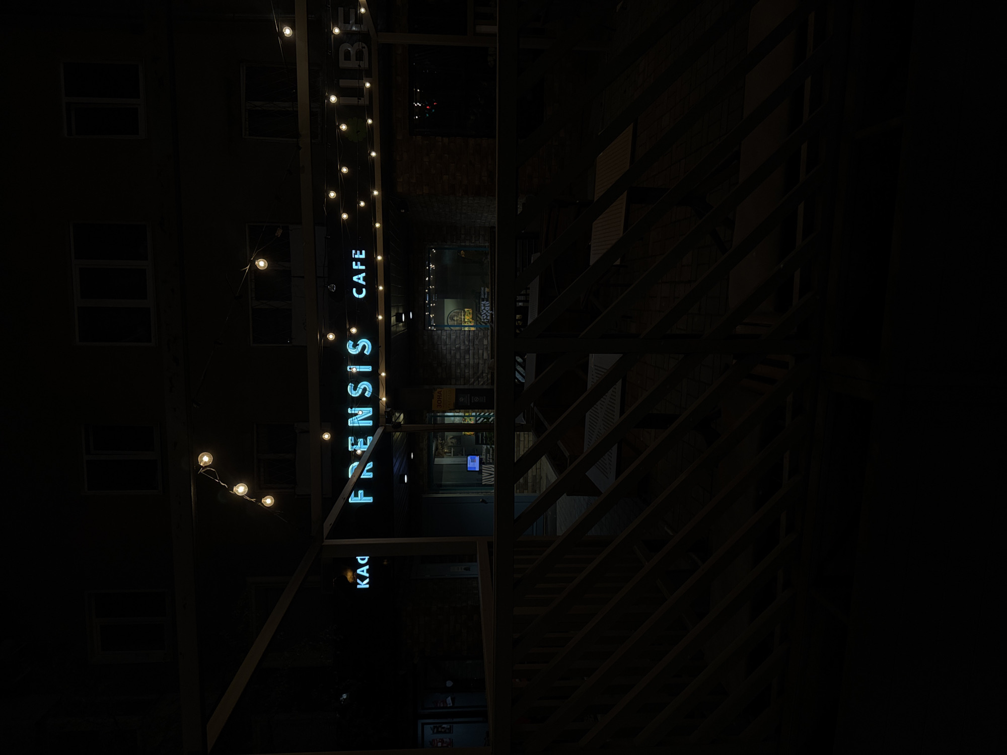 The image shows the Frensis cafe located in Izhevsk, Russia. The blue neon sign "FRENSIS CAFE" is vertically oriented and illuminated. String lights are visible nearby. The facade of a building with windows can be seen behind the sign. A wooden structure with stairs and diagonal supports dominates the right side of the image. The overall lighting is dim, suggesting it is nighttime.