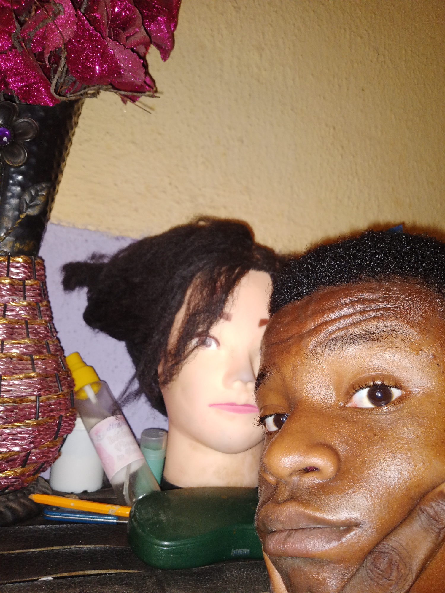 The image depicts a close-up selfie of a young man with dark skin and short, dark hair. His face is the primary subject, with his left hand partially visible, supporting his chin. His expression is neutral.
Behind him is a mannequin head with black hair. A decorative vase filled with artificial pink flowers is located to the left of the mannequin. On a surface in front of the mannequin and vase are various items, including a spray bottle, other small bottles, pens, and a green glasses case.
The background features a wall with a two-tone color scheme, with a light beige color on the upper portion and a lilac color below.
The location is Narayi, Nigeria.