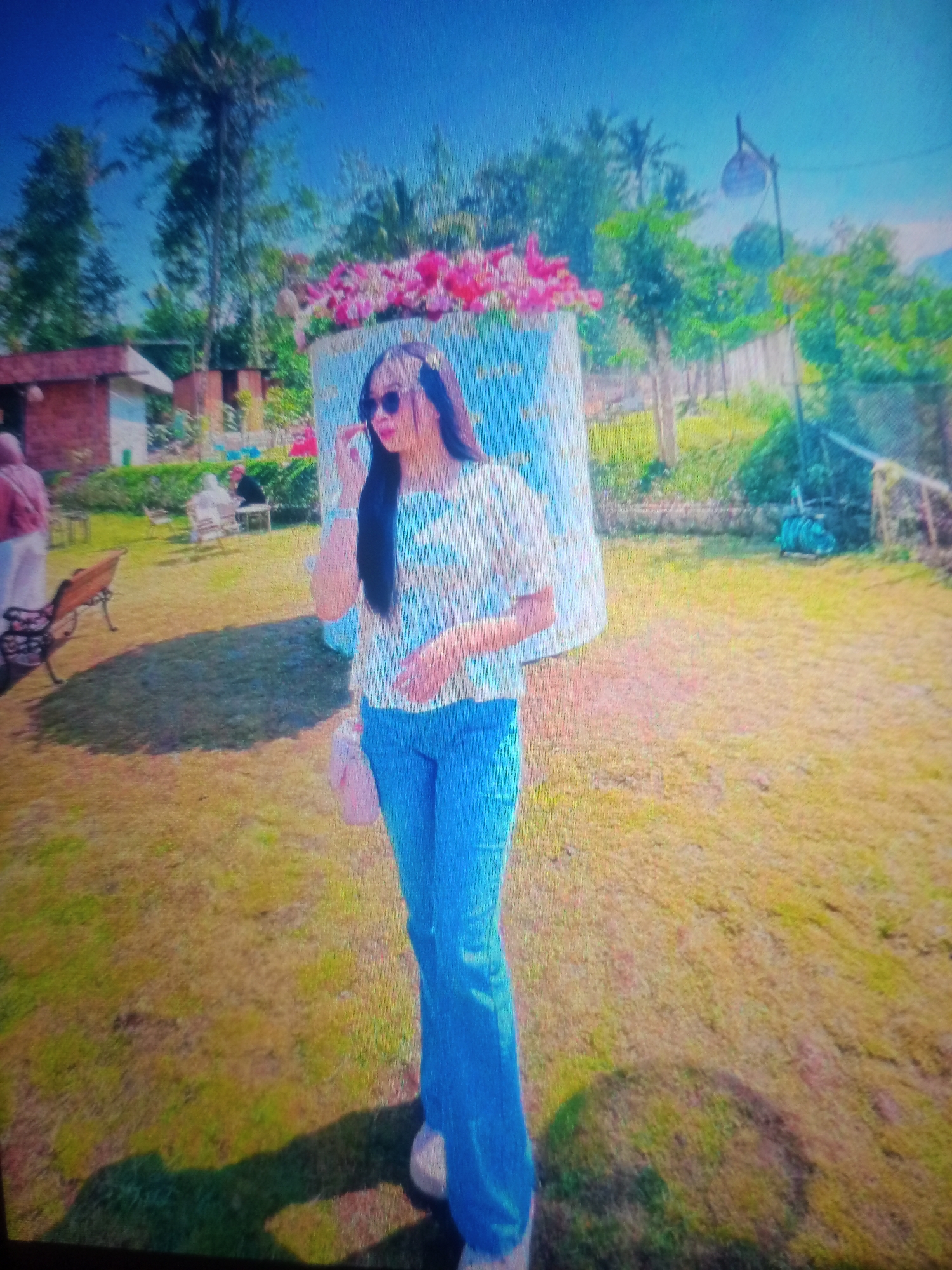A woman stands in a grassy outdoor area in Nusa Agung, Indonesia. She has long dark hair and is wearing sunglasses, a light-colored patterned shirt, and blue flared jeans. She has a small pink purse and is touching her hair. Behind her, a large floral arrangement sits atop a blue and white structure. In the background, there are trees, a brick building, a bench, and other people seated at tables. The sky is bright blue.