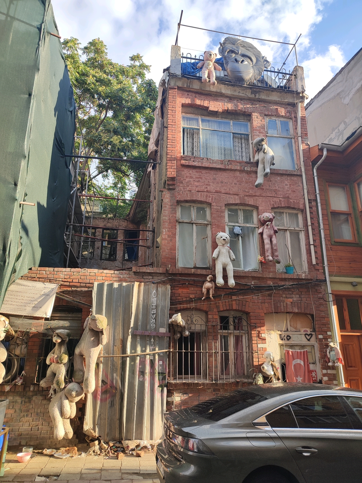 The image shows a dilapidated brick building in Kadıköy, Turkey, adorned with numerous large stuffed teddy bears hanging from windows, balconies, and scaffolding. The bears, varying in size and color, are positioned in a whimsical, almost haunting manner, some appearing to be in mid-fall. The building itself exhibits signs of neglect, with peeling paint, exposed brickwork, and some windows covered with metal bars.
On the rooftop, a large, gray, sculpted gorilla head is prominently displayed, casting a surreal shadow. A small teddy bear is also perched near the gorilla head. Other details include a Turkish flag hanging from a window, a mannequin head with a brown garment draped around it, and a dark-colored sedan parked in front of the building. The overall atmosphere is one of urban decay punctuated by quirky and unsettling artistic expression. The surrounding environment appears to be a narrow street with other buildings visible, suggesting a densely populated urban area. The sky is partly cloudy.
