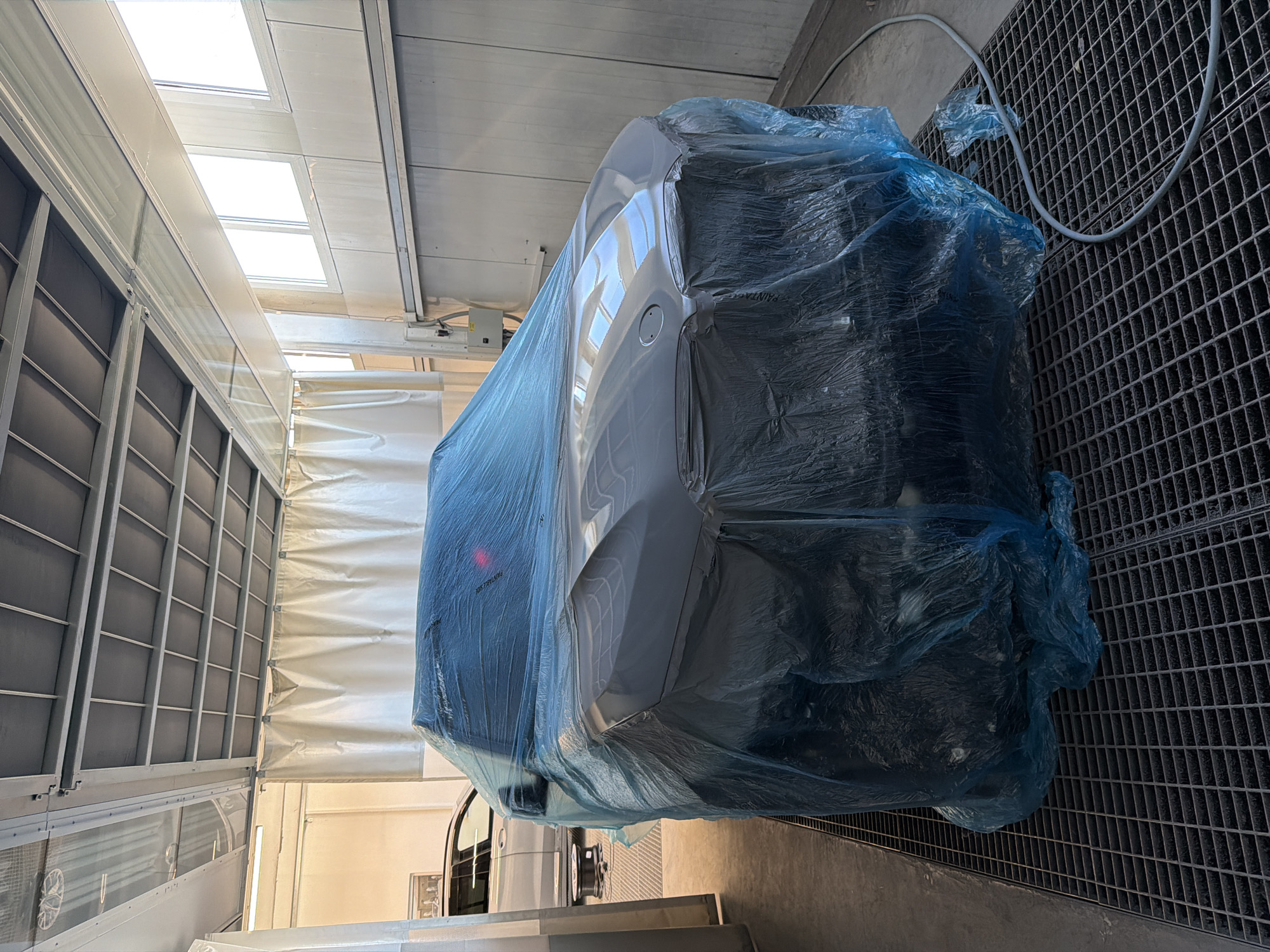 The image shows a light-colored car, possibly white or silver, largely covered by a translucent blue plastic sheet within what appears to be an automotive workshop or paint booth. The car is positioned on a floor composed of dark metal grates, which often serve for ventilation or drainage in such facilities. A light-colored hose or cable is visible snaking across the grated floor.
The setting is an indoor environment, brightly lit by natural light streaming through multiple rectangular skylights in the ceiling, indicating it is daytime. The interior walls feature large, light-colored panels, with some sections displaying horizontal divisions or framing. To the left, a structured wall with vertical and horizontal frames is visible, while to the right, a soft, white, possibly flexible curtain-like wall can be seen.
The blue plastic sheeting completely envelops the car, suggesting protection during or after a process like painting or detailing. A faint, possibly partial, text "PAINTAG" or similar is visible on the blue plastic covering the car. No individuals are present in the scene, but the professional setup implies an automotive service activity is either underway or has recently been completed.
