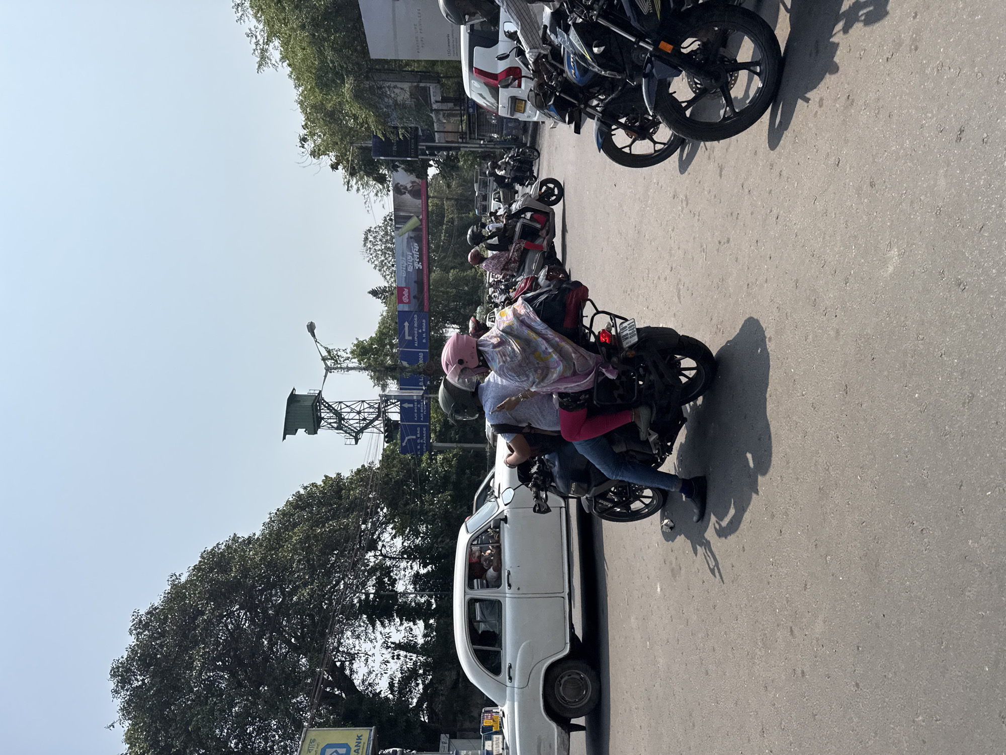 The image depicts a street scene in Kolkata, India. A white Hindustan Ambassador taxi car is positioned on the street alongside several parked motorcycles. A motorcycle with two individuals is in the foreground: the driver wearing a helmet and the passenger, a child also wearing a helmet, with a pink tint, and a colorful patterned wrap or shawl. Several other motorcycles and a white van or truck are parked behind the taxi. In the background, utility poles and trees line the street, with billboards and signs visible above. Road signage indicates Alipore Road and D.H. Road. The sky is clear and bright.