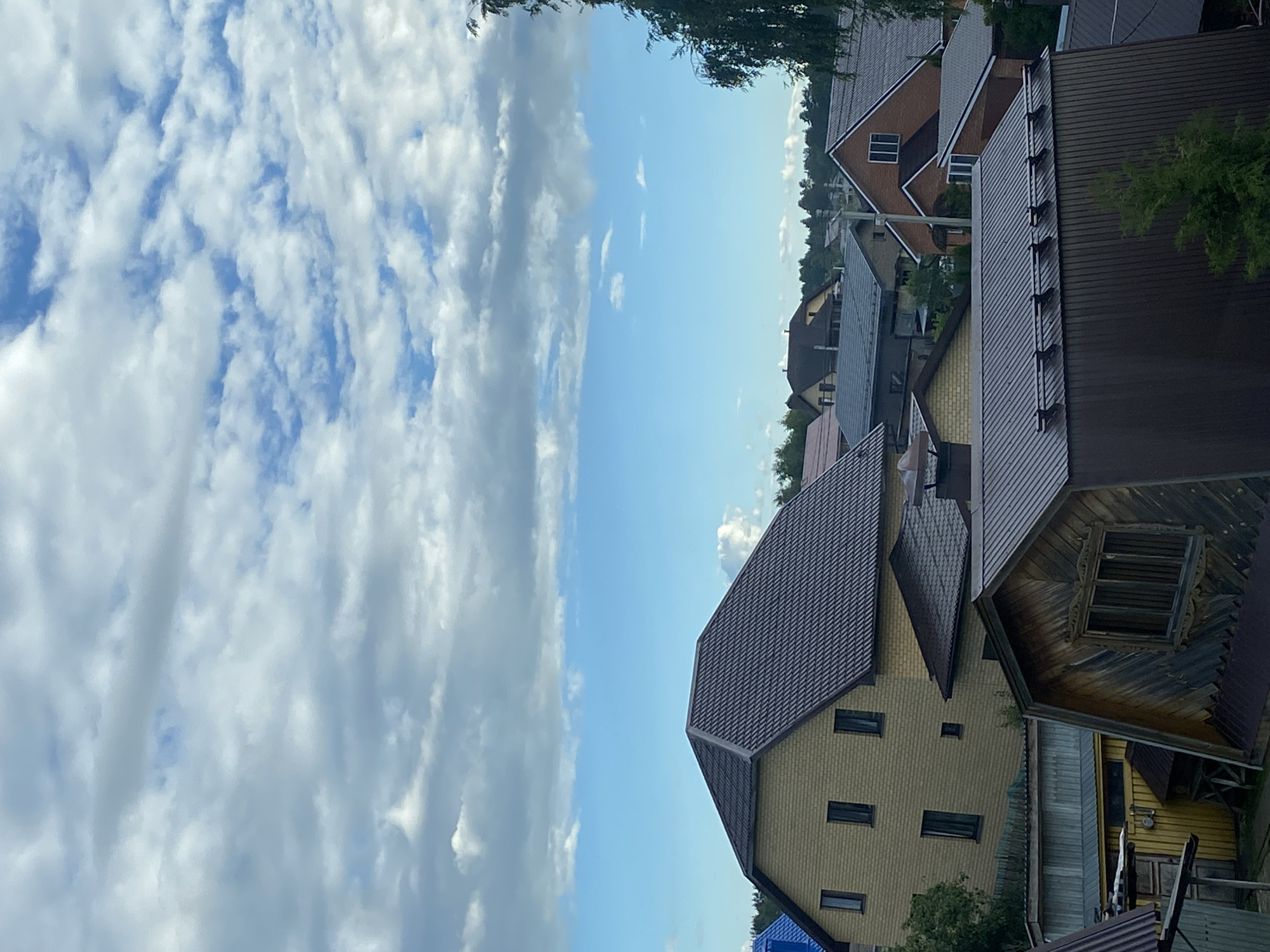 The image depicts a residential area with several houses and a partly cloudy sky. The perspective is from a slightly elevated position, looking out over rooftops. The houses have various architectural styles and roofing materials, including dark gray and brown shingles, and some feature visible windows. One house in the foreground is a light yellow color with multiple rectangular windows. Another house to the right has a wooden facade with an ornate window frame and visible wooden beams underneath the roof. Trees and foliage are visible interspersed between the buildings, suggesting a suburban or rural setting. The sky is a mixture of blue sky and white, puffy clouds, indicating daytime, likely late afternoon or early evening due to the lighting. The overall impression is one of a quiet, established neighborhood. No people are visible in the frame. There is no visible text in the image. The location context is Zelenodol'sk, Russia.