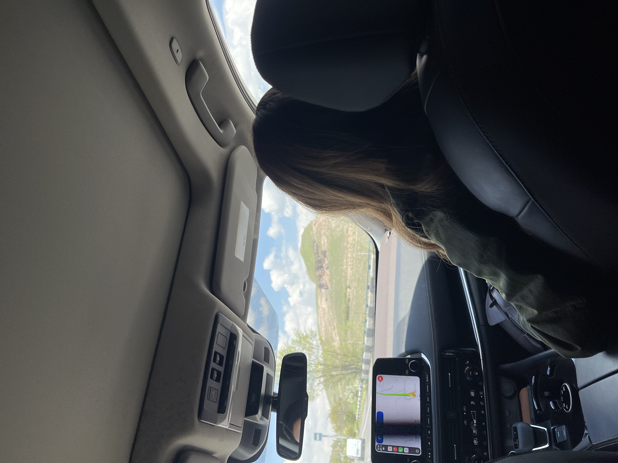 The photograph was taken from inside a vehicle, likely a car, while traveling on a road near Arzni, Armenia, based on the landscape visible through the windshield. A person with long brown hair is in the passenger seat, their head near the headrest. The vehicle's interior features a rearview mirror, a display screen showing a navigation app, and various control panels. Outside the vehicle, the scenery includes a green hillside and a cloudy sky.