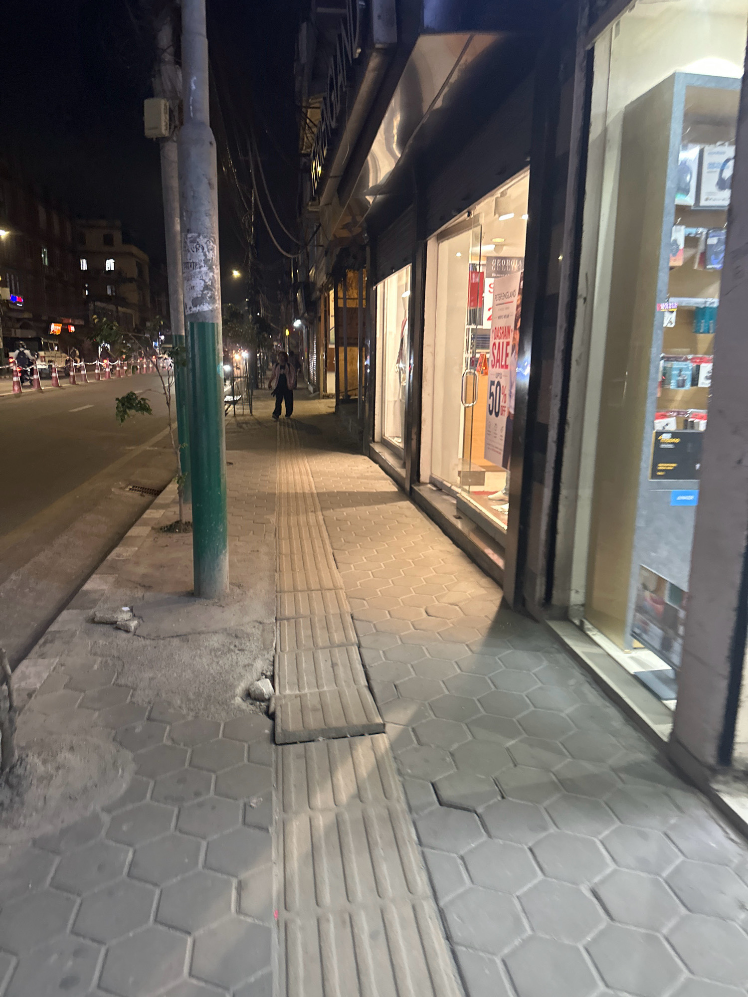 The image shows a street scene at night in Kathmandu Metropolitan City, Nepal. The left side of the image features a sidewalk with hexagonal paving stones, a metal pole, and a street with traffic cones and vehicles. The right side of the image displays the storefront of a shop with a large window showcasing merchandise and a prominent "SALE 50%" sign. A person is walking on the sidewalk in the background. The overall environment is urban and appears to be a commercial area.