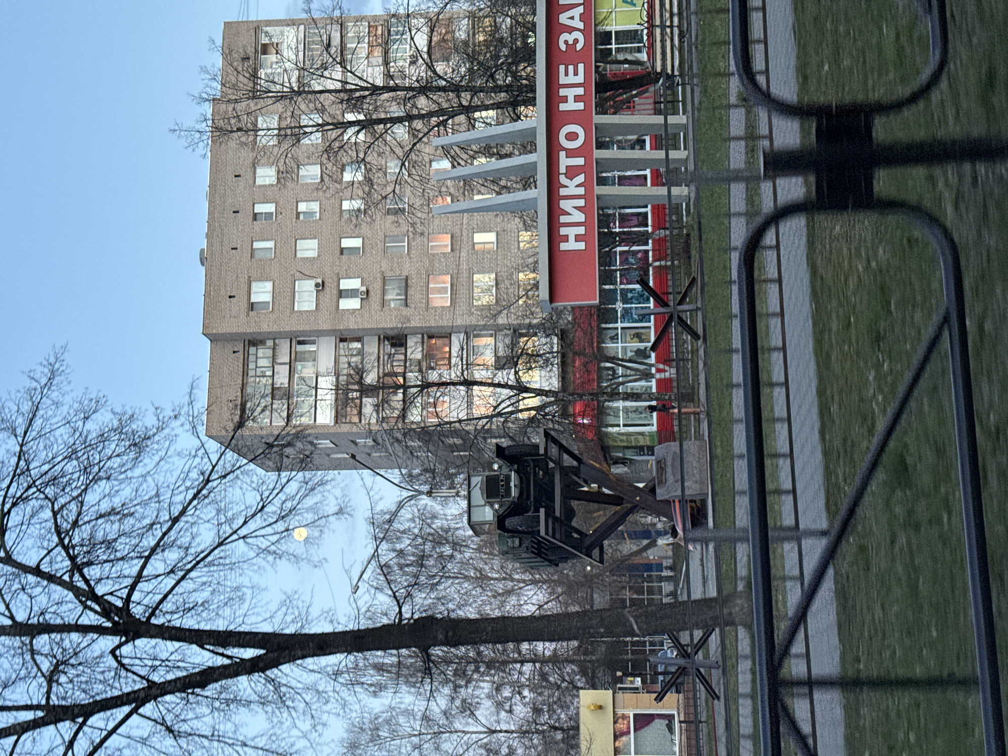 The image, rotated 90 degrees clockwise, depicts an urban scene featuring a tall, multi-story building constructed of light brown brick. Bare tree branches, suggesting late fall or winter, frame the view against a clear, light blue sky. A bright, circular object, likely the moon, is visible in the sky through the branches.
In the midground, a prominent red sign with white Cyrillic letters reads "НИКТО НЕ ЗАБ". This is part of the common Russian phrase "Никто не забыт" (Nikto ne zabyt), meaning "No one is forgotten," often associated with war memorials. Below and in front of this sign, a dark, heavy-duty vehicle, resembling a military truck or a monument thereof, is displayed on a raised structure. Several dark, metal anti-tank obstacles, known as "Hedgehogs," are visible on the ground near the vehicle and in the immediate foreground, reinforcing a memorial or historical theme.
A dark metal fence runs diagonally across the foreground, separating a grassy area from a paved pathway. The building itself has numerous windows, some of which show interior lights, suggesting it is a residential or mixed-use commercial building. The overall lighting indicates either early morning or late afternoon. A small kiosk-like structure is partially visible in the lower right, with a person seen through its window. The scene strongly suggests a location in an Eastern European country, likely Russia or Ukraine, given the text and memorial elements.
